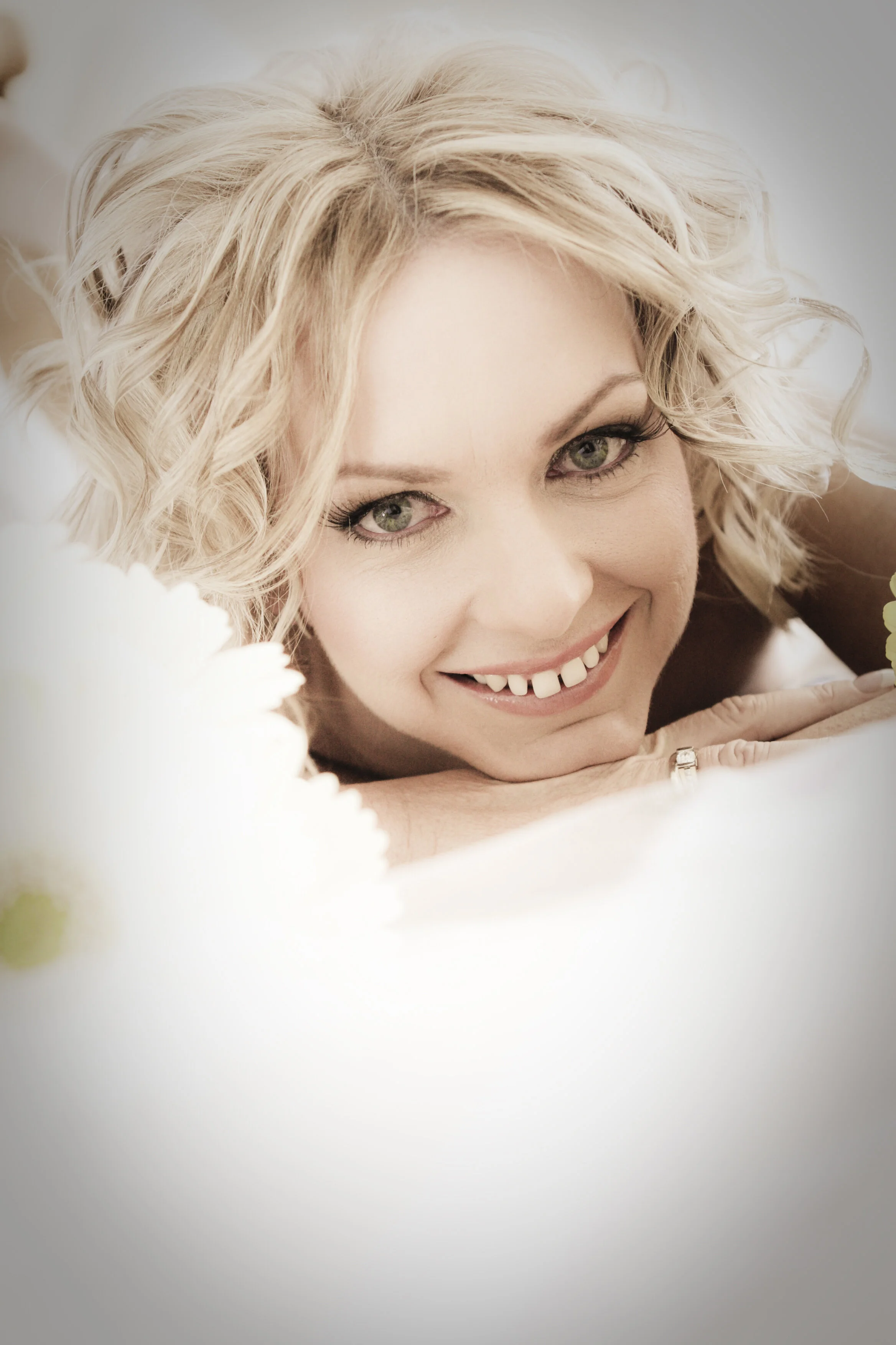 Close-up of a smiling woman with blonde, curly hair, lying down and resting her head on her arms, surrounded by soft white flowers.