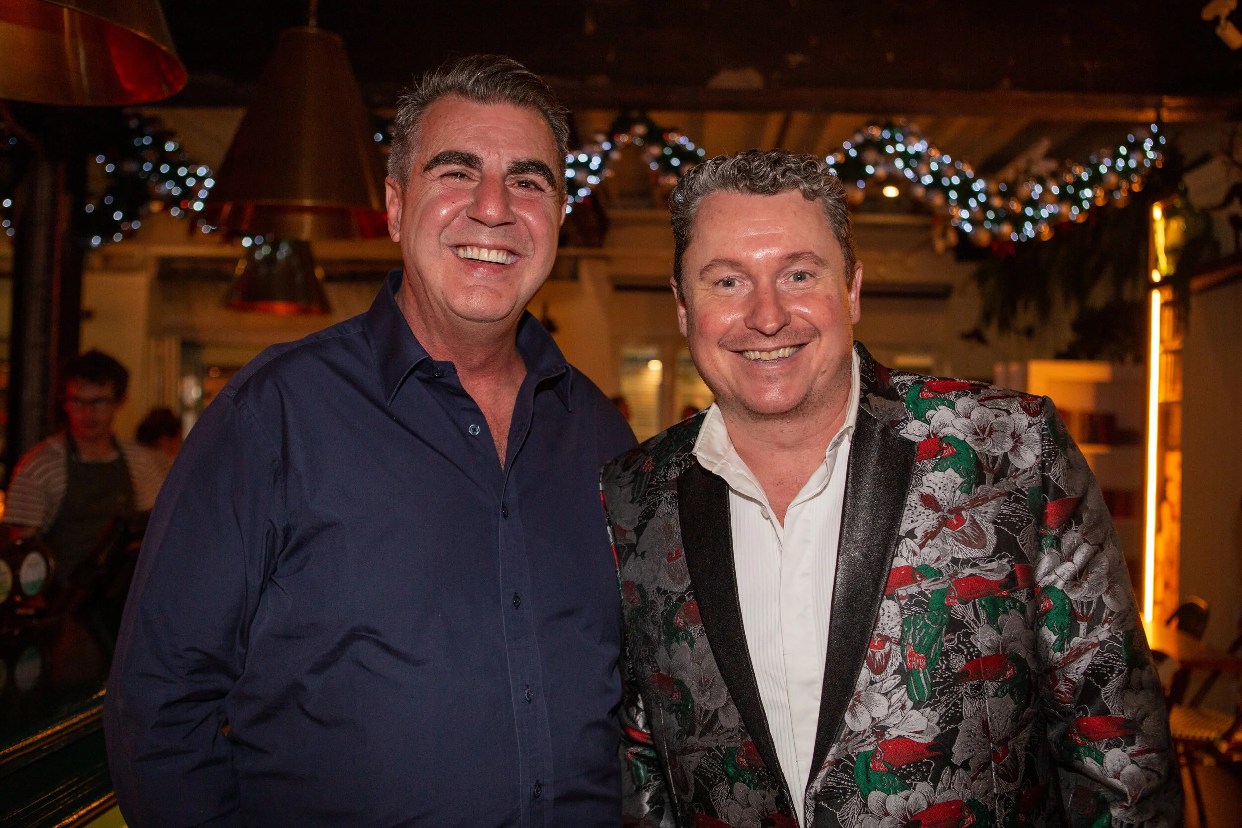 Two smiling men at a festive indoor event, one in a dark blue shirt and the other in a floral blazer with parrot and flower patterns, with holiday decorations and a person in the background.