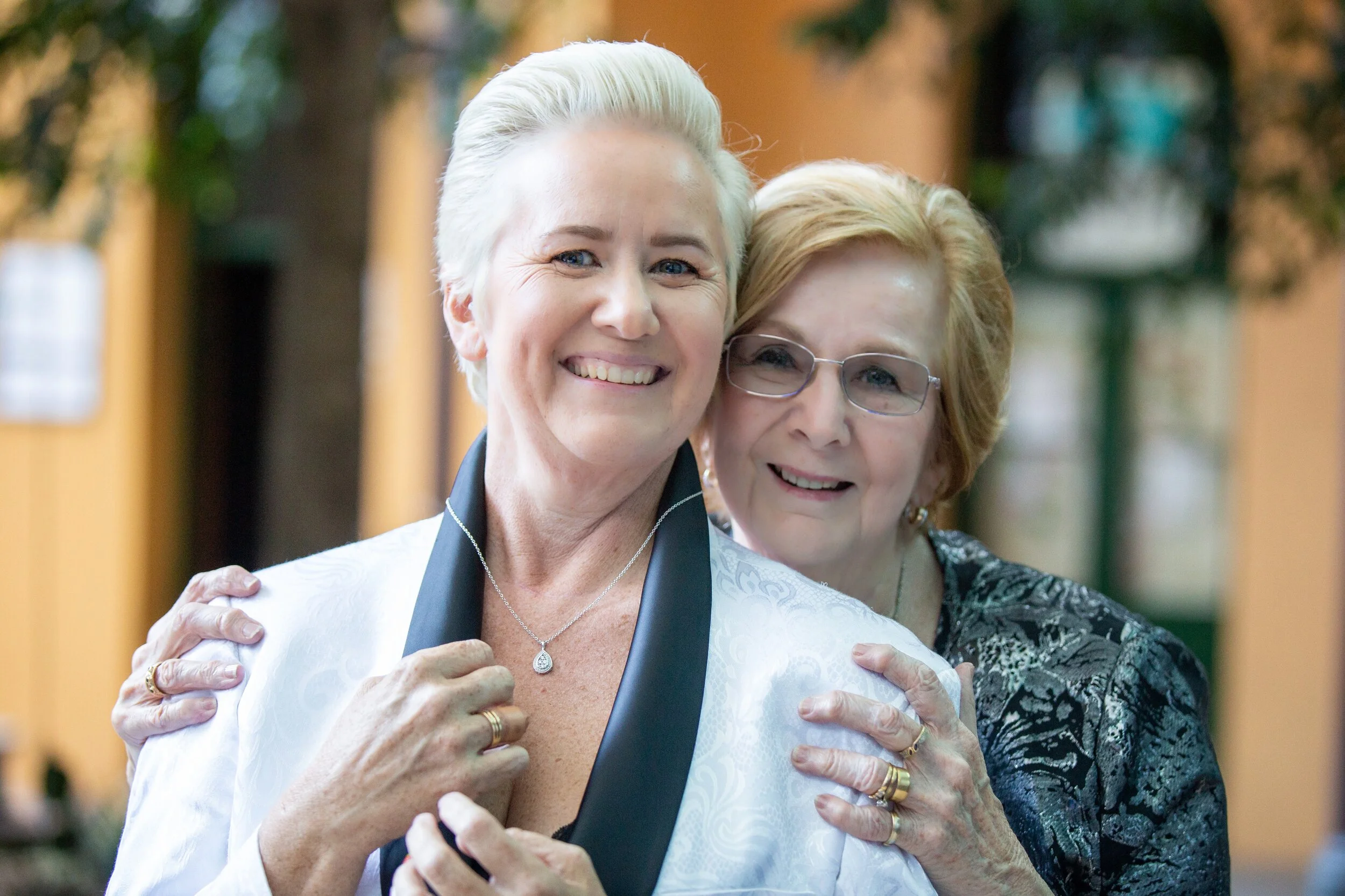 Two smiling women, one with short white hair and the other with short red hair and glasses, sharing a happy moment together indoors.