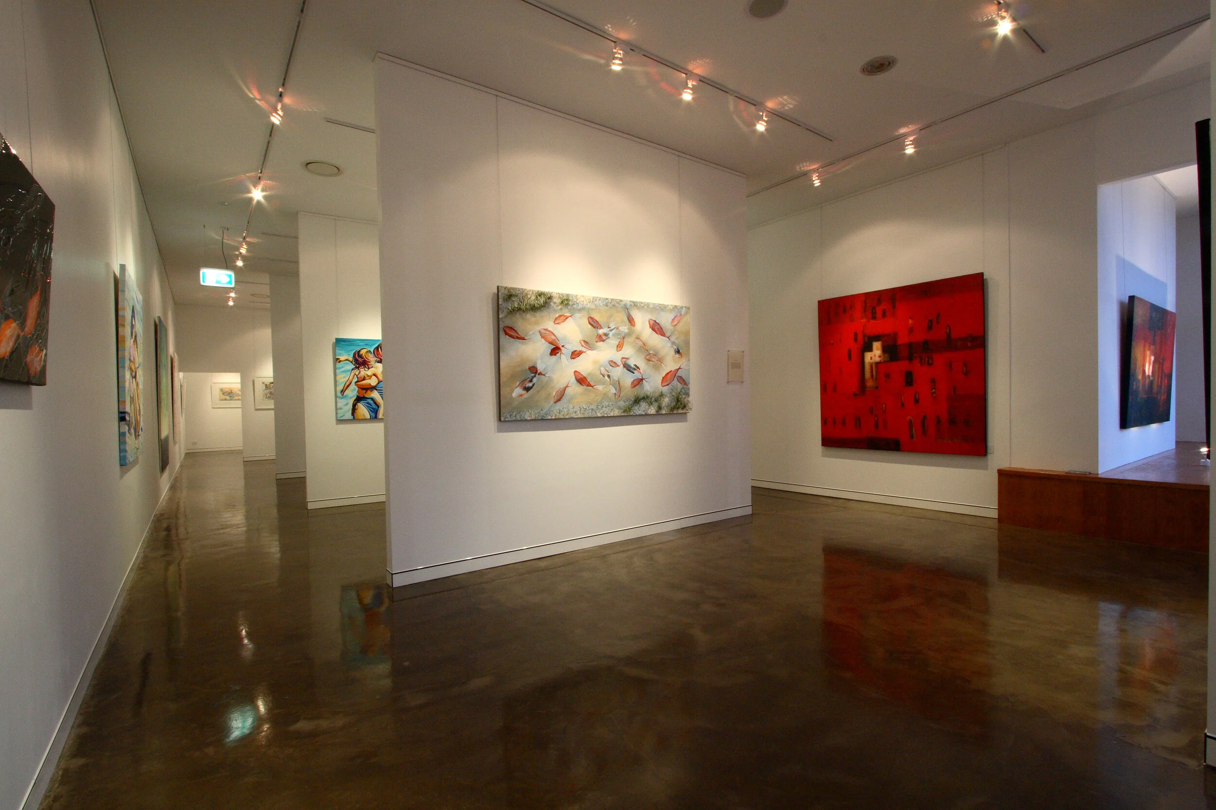 Art gallery with white walls displaying colorful paintings, including a red abstract piece and a painting of swirling leaves, with wooden flooring and ceiling track lighting.