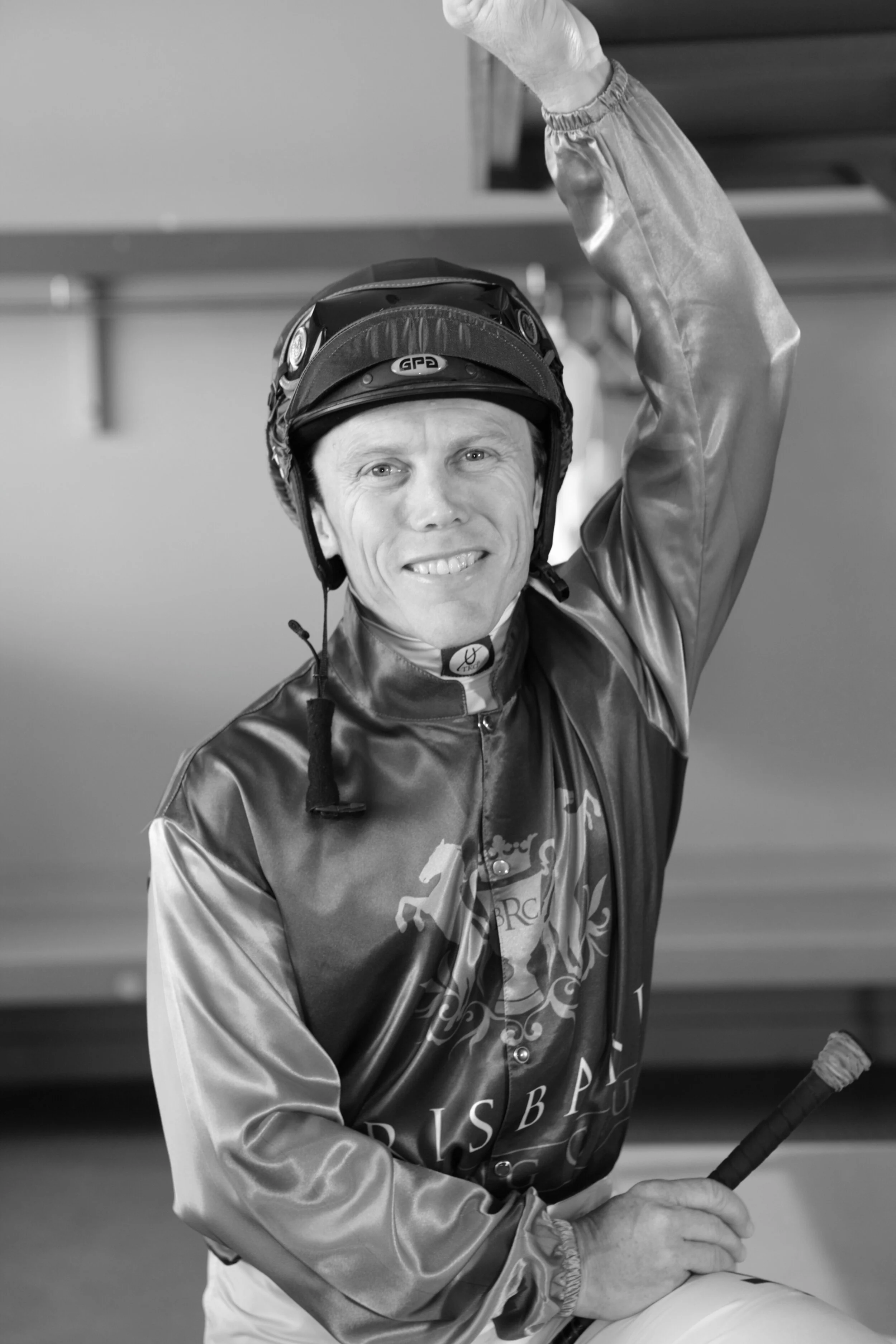 A smiling male jockey in racing gear and helmet, holding a riding crop, inside a stable.