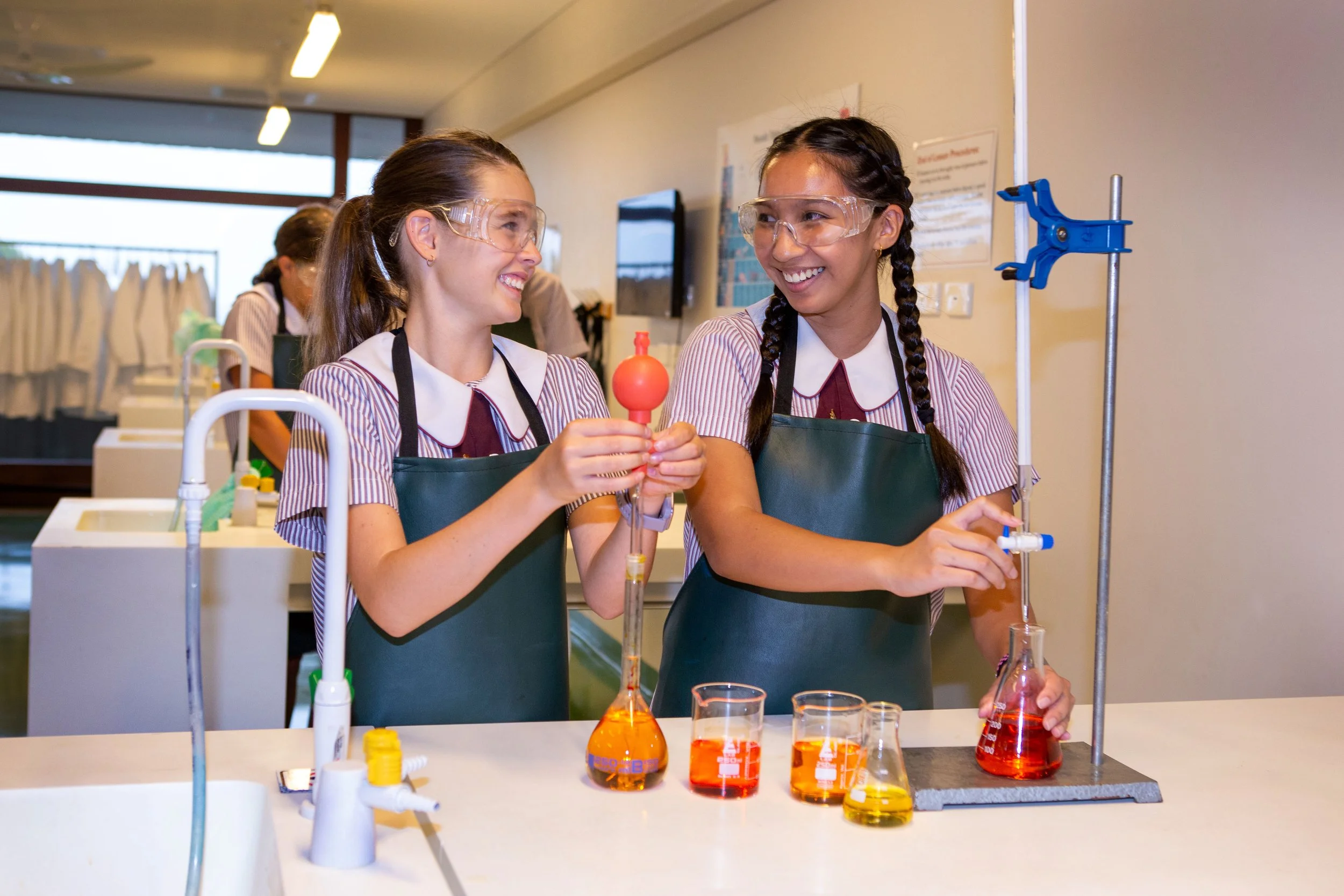 Two young girls in a science classroom conducting an experiment with colorful liquids in glassware, smiling and wearing safety goggles and aprons.