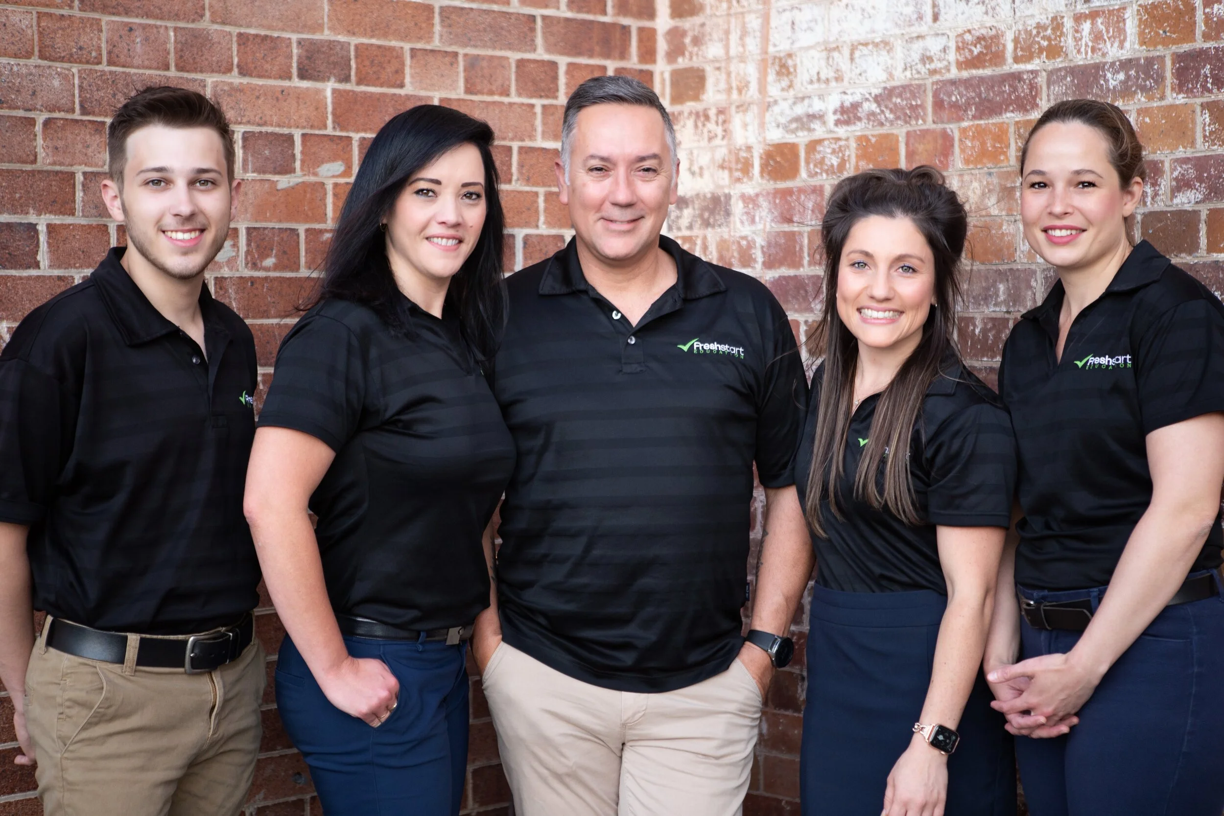 Group of five people standing in front of a brick wall, all wearing matching black shirts with a logo.