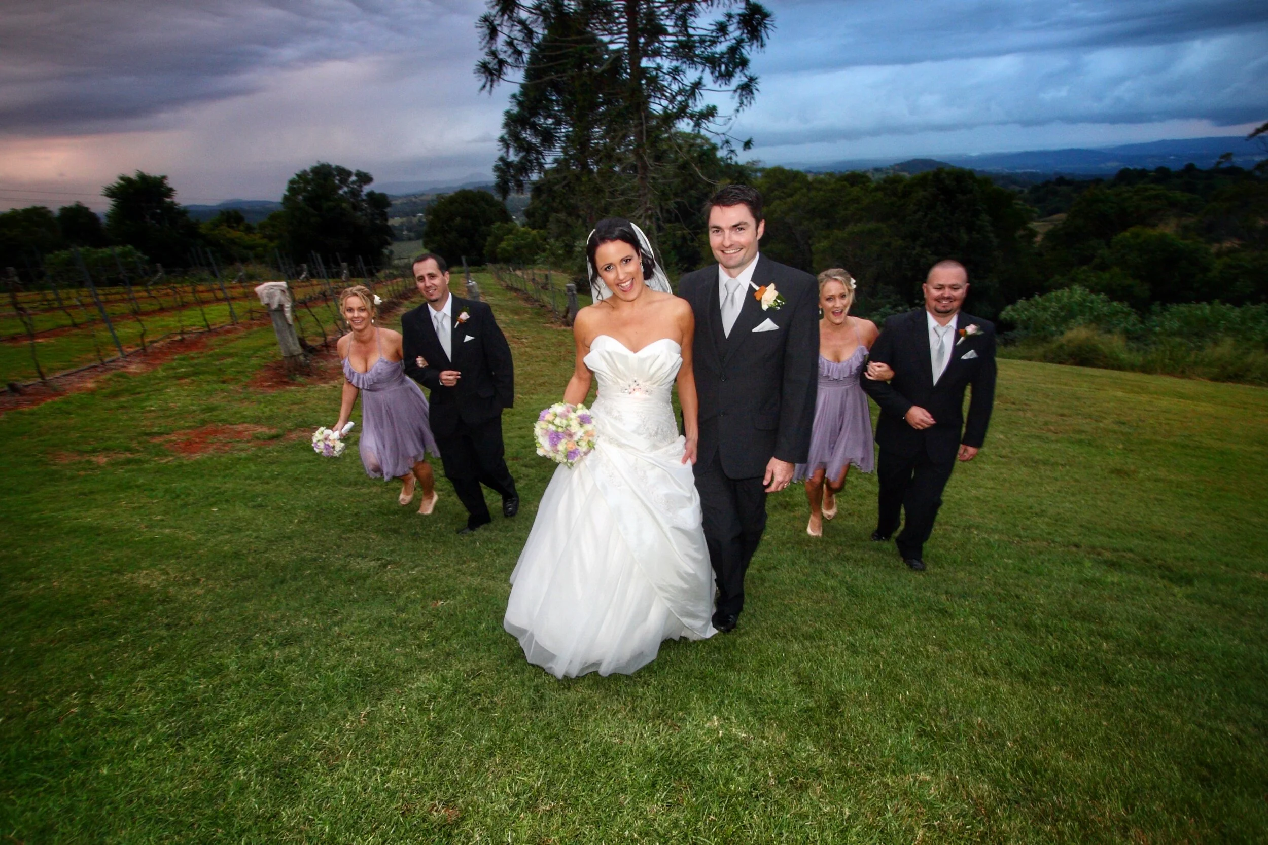A bride and groom walking on a grassy field with friends in the background, vineyard in the distance, during cloudy weather.