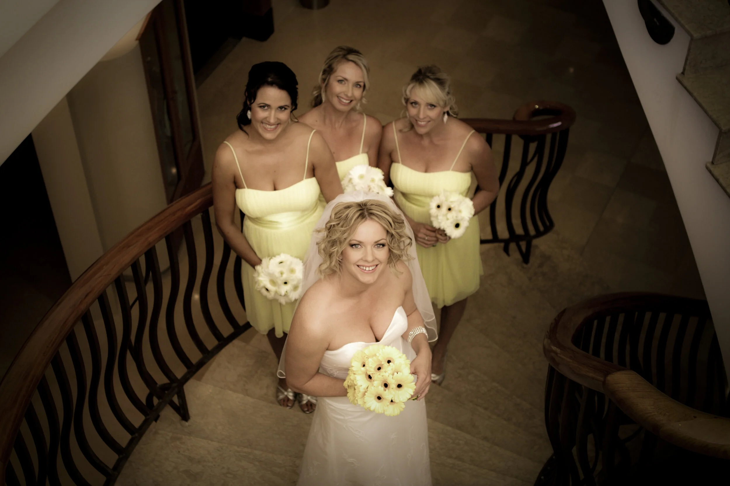 Wedding bride in white dress holding a bouquet, surrounded by four bridesmaids in yellow dresses, standing on a staircase.