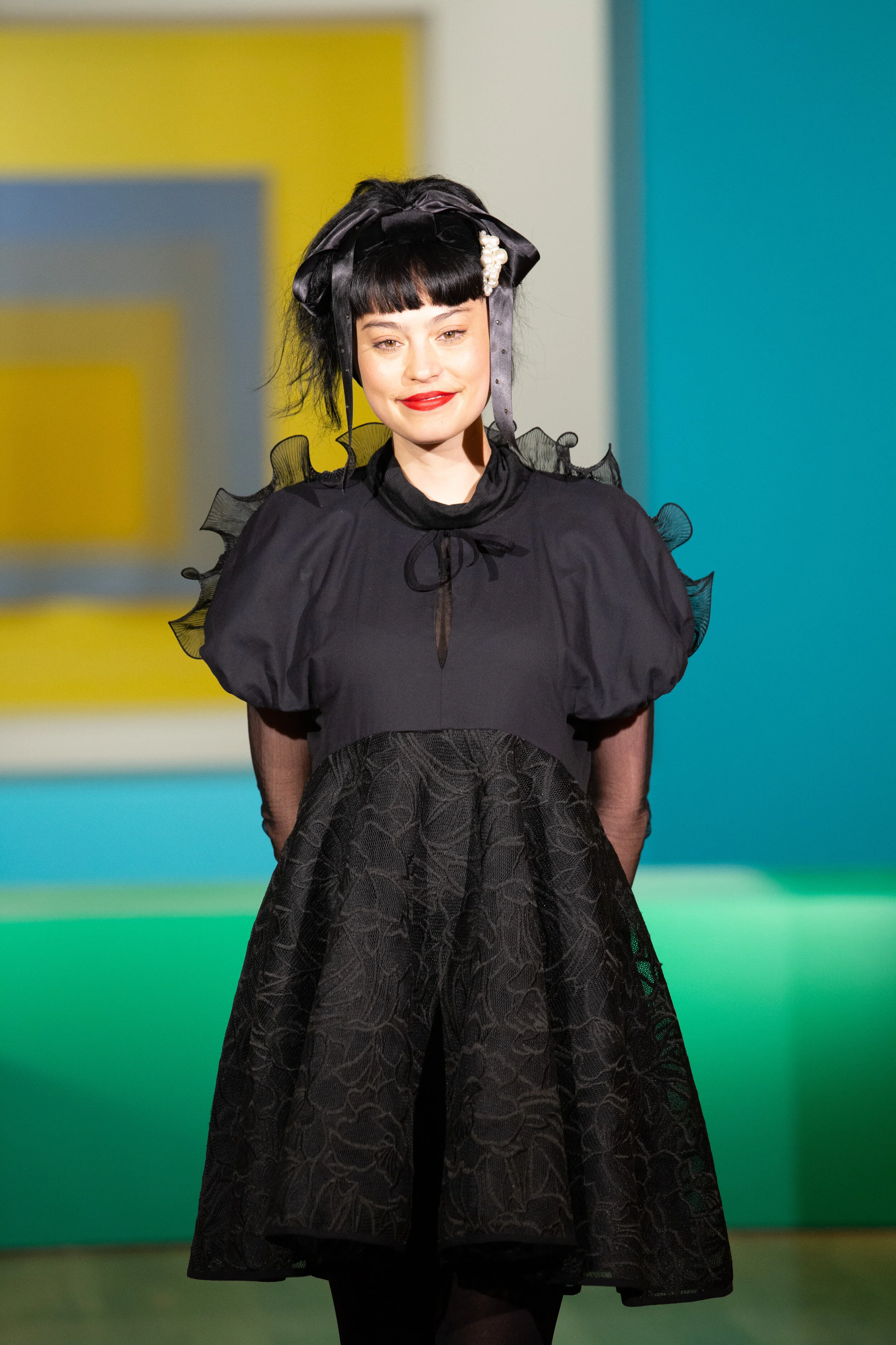 Woman with short black hair, bangs, wearing a black dress with puffed sleeves and lace overlay, standing in front of colorful background.