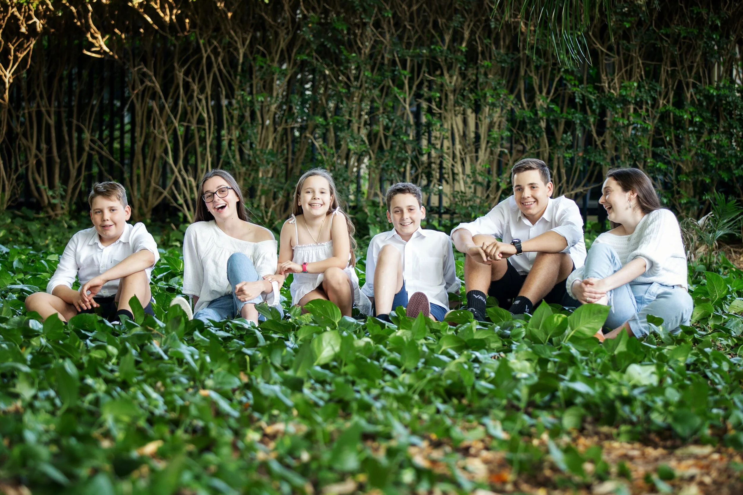 Six children sitting on green ground cover plants in a wooded area, smiling and enjoying each other's company.
