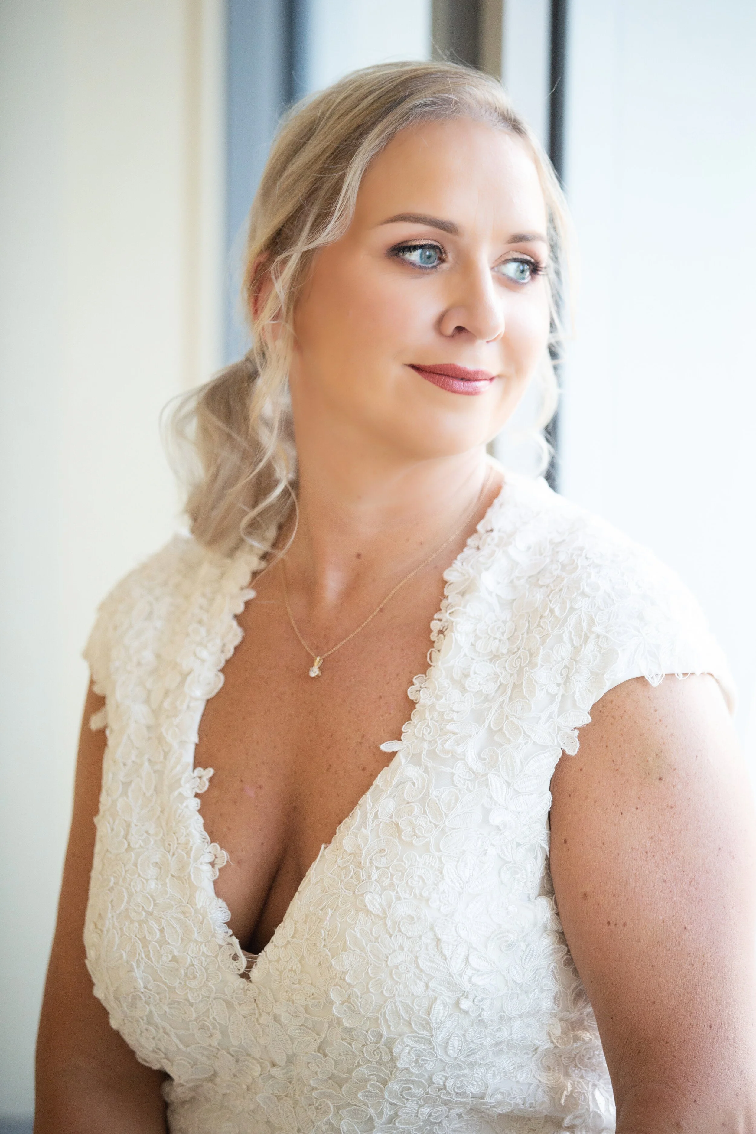 A woman with blonde hair styled in loose waves, wearing a white lace dress with floral detailing, looking out the window with a gentle smile.