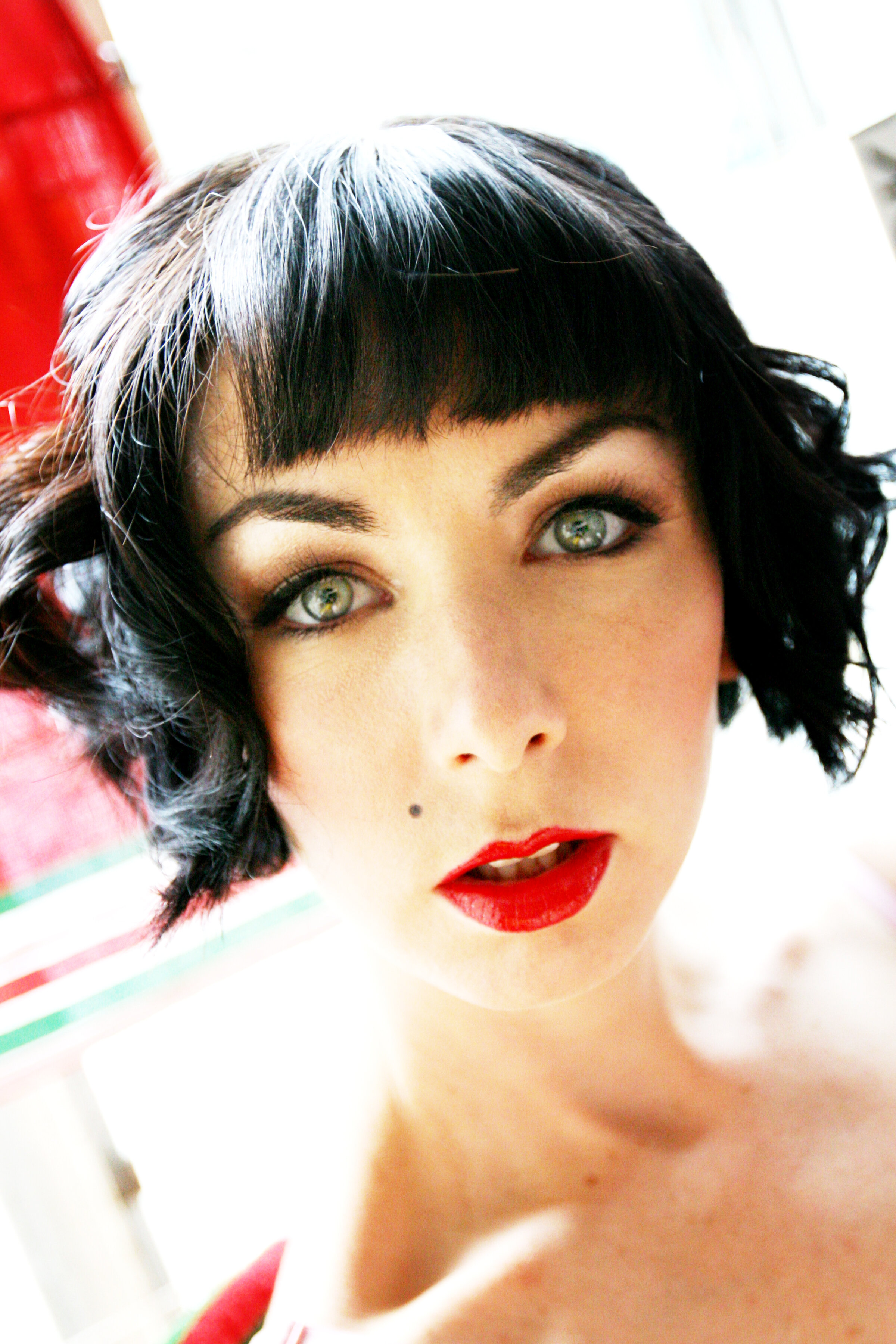Close-up portrait of a woman with short black hair, green eyes, bold red lipstick, and a beauty mark on her left cheek, looking directly at the camera.