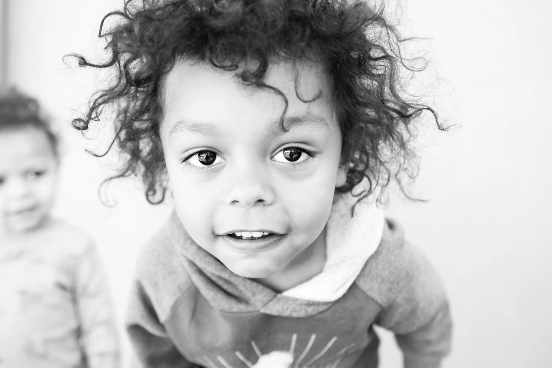 Close-up of a young child with curly hair looking directly at the camera, smiling, with another child in the background.