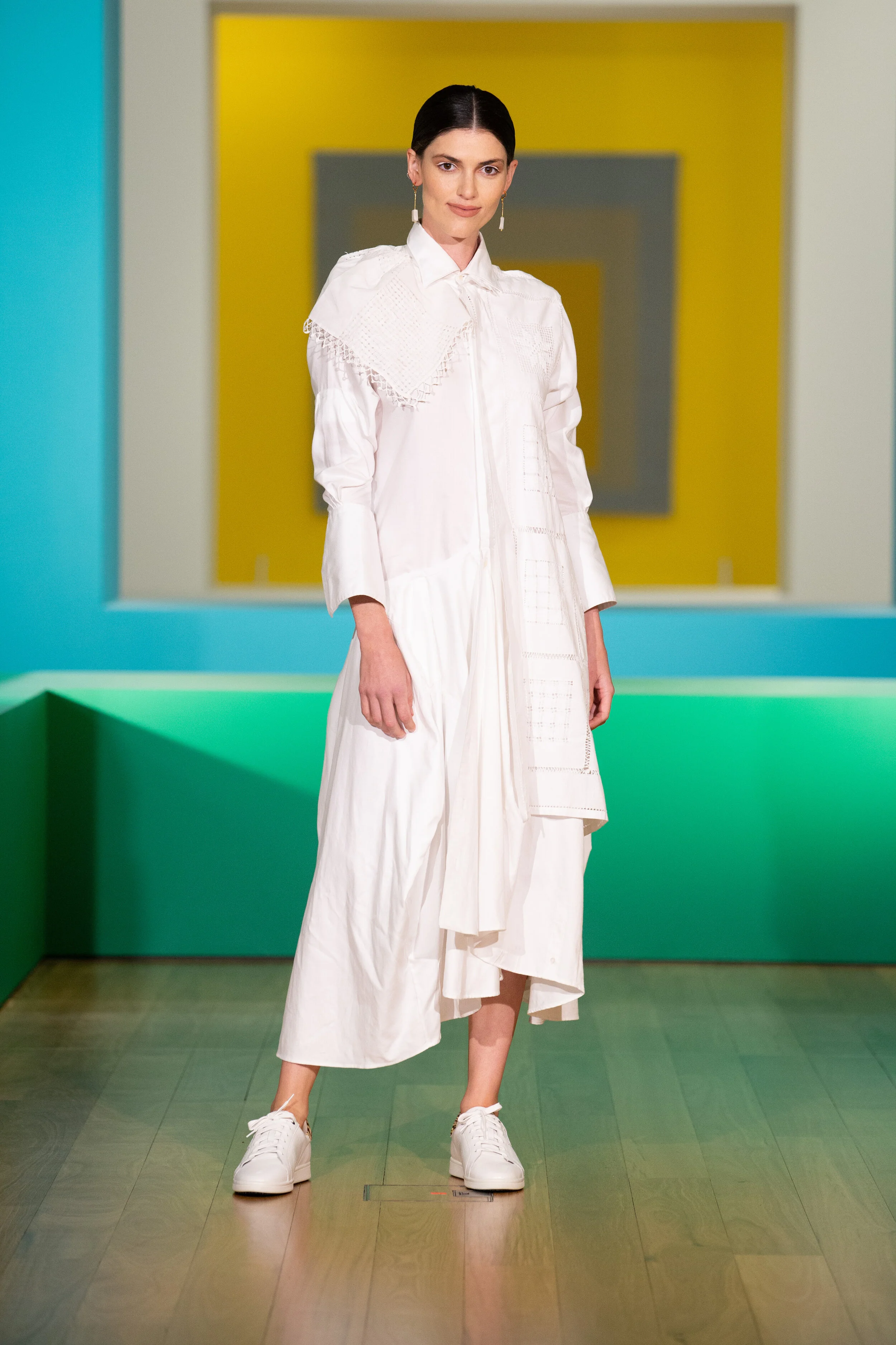 A woman with dark hair in a white oversized shirt and matching wide-leg pants, wearing white sneakers, standing in front of colorful geometric background.