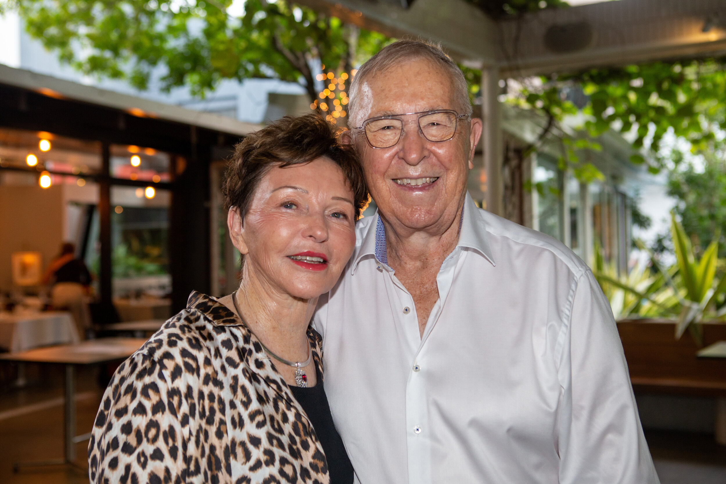 An elderly couple smiling and posing together indoors with green plants and string lights in the background.