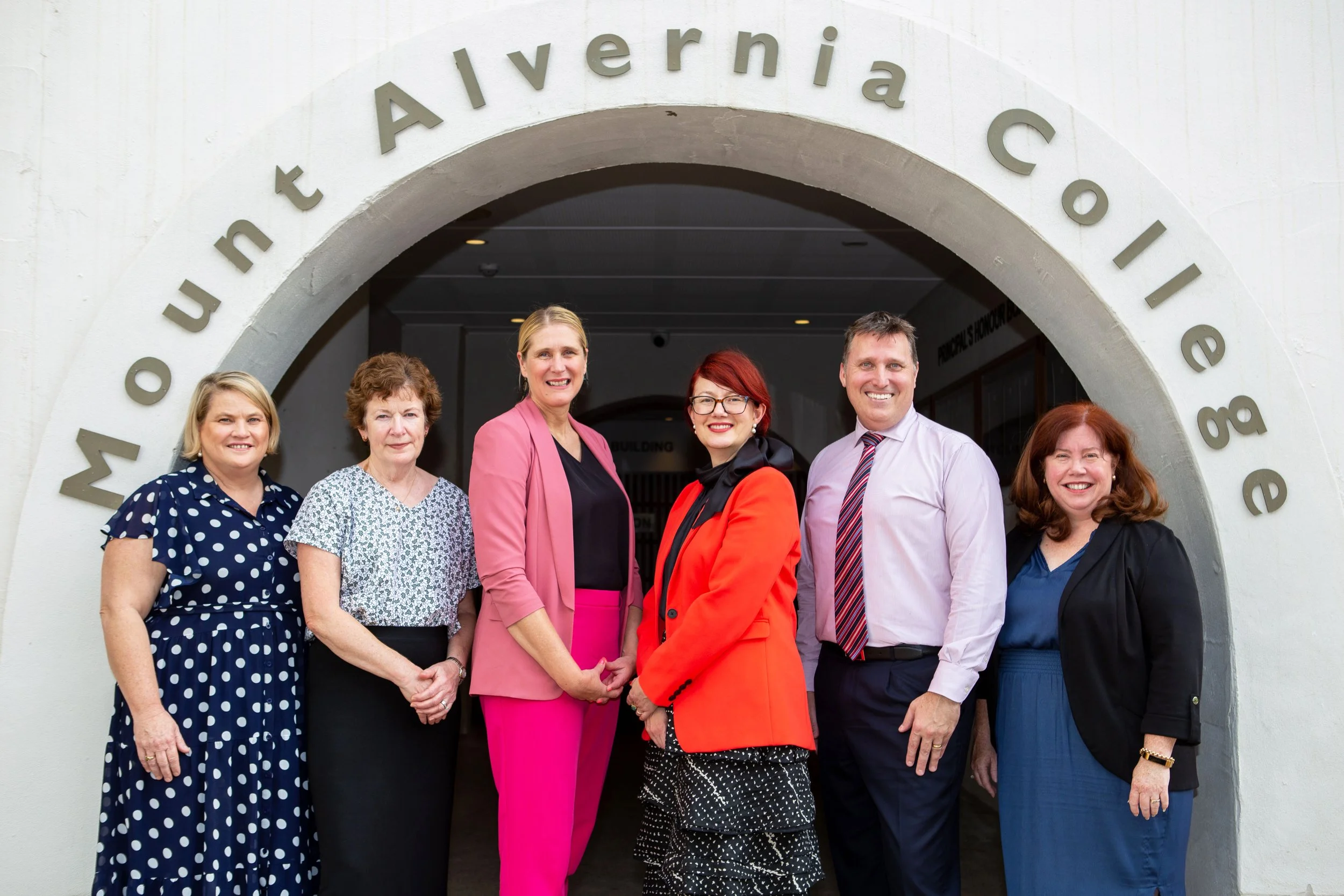 Group of six diverse professionals standing in front of a building with an arch that says "Mont Alvernia College"
