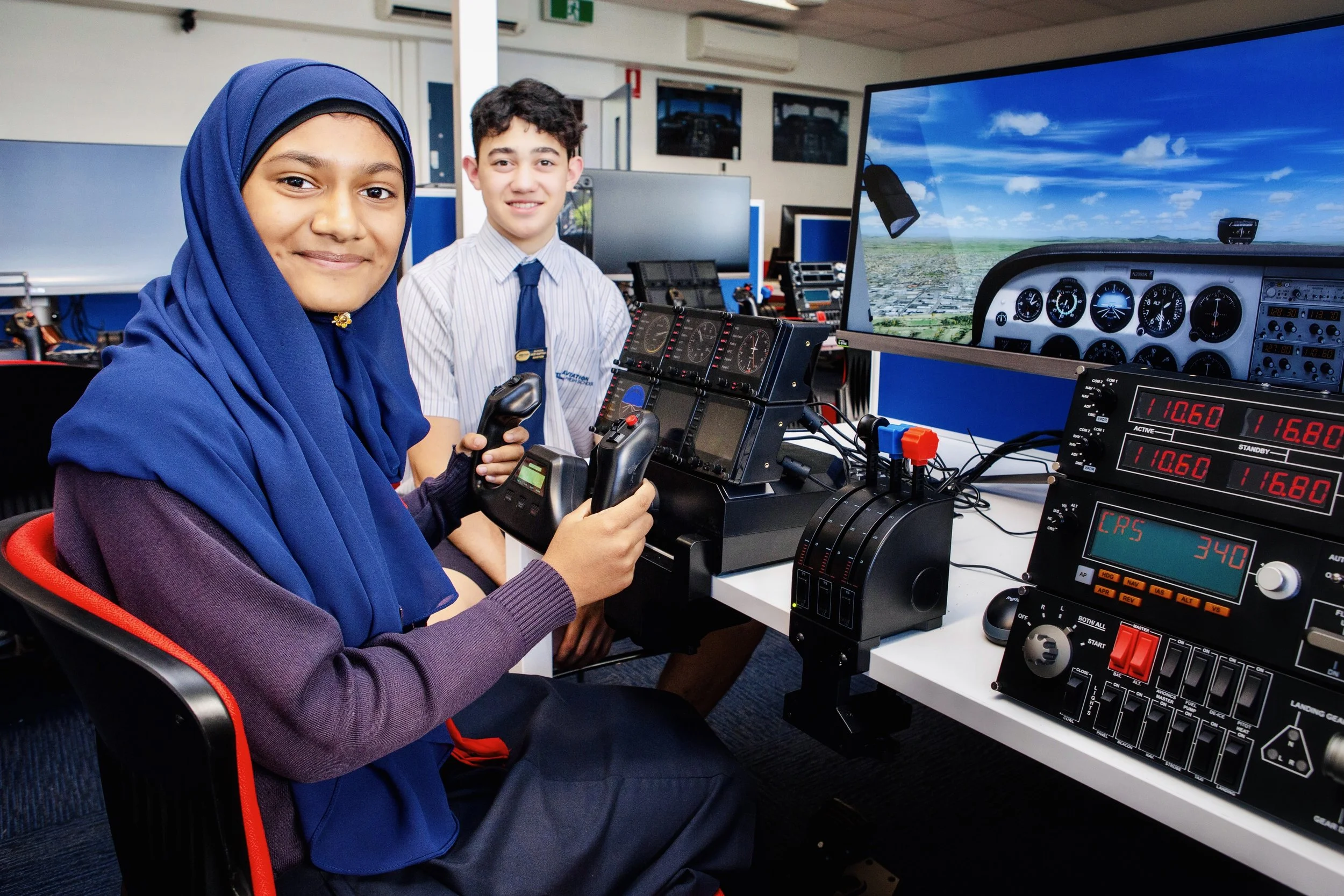 Two students in a flight simulator, one girl wearing a blue hijab and a girl beside her, piloting a simulated airplane cockpit with controls and a large screen showing the sky.