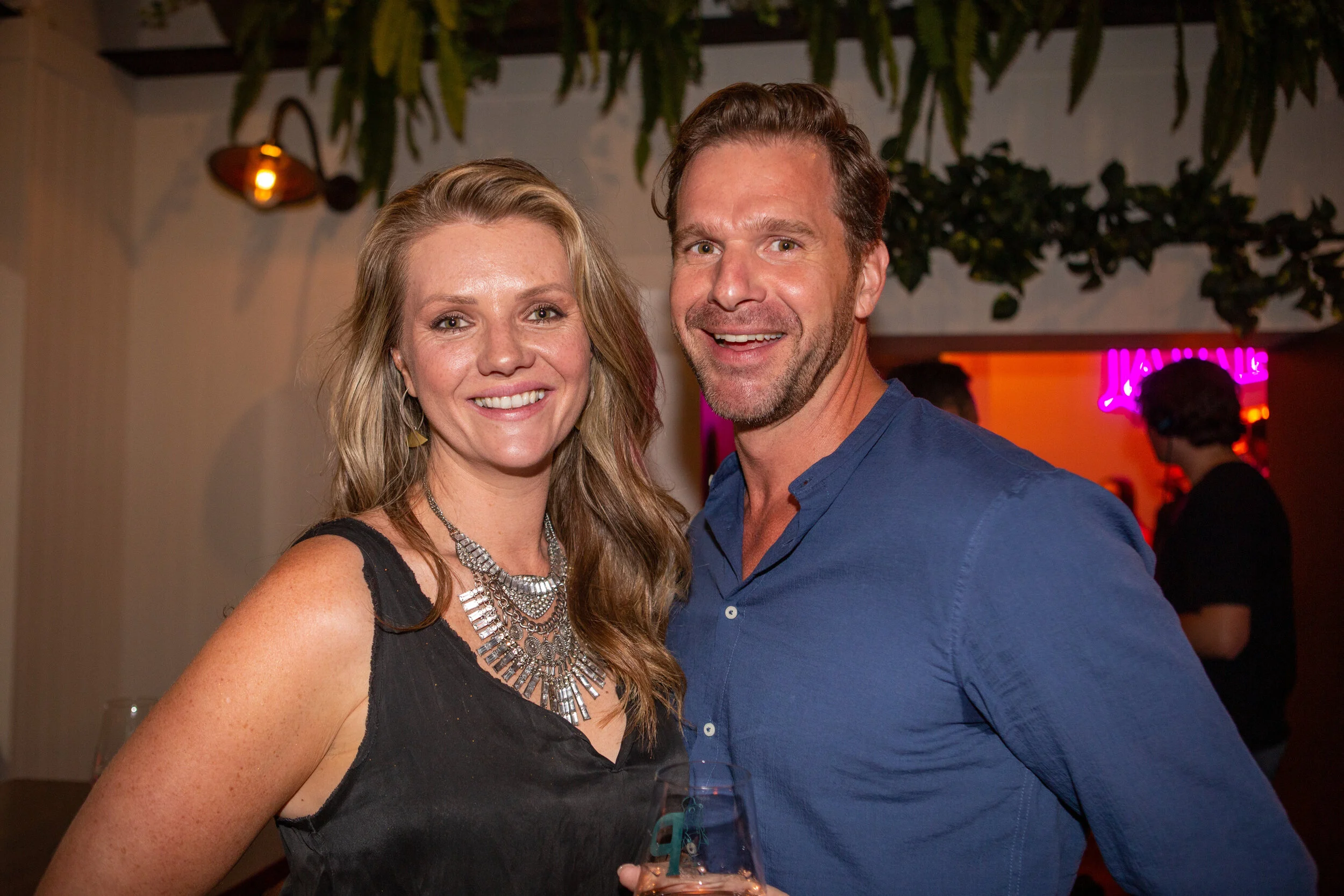 A smiling man and woman at a social event, with the woman wearing a black dress and a layered silver necklace, and the man wearing a blue shirt, holding a glass of wine.