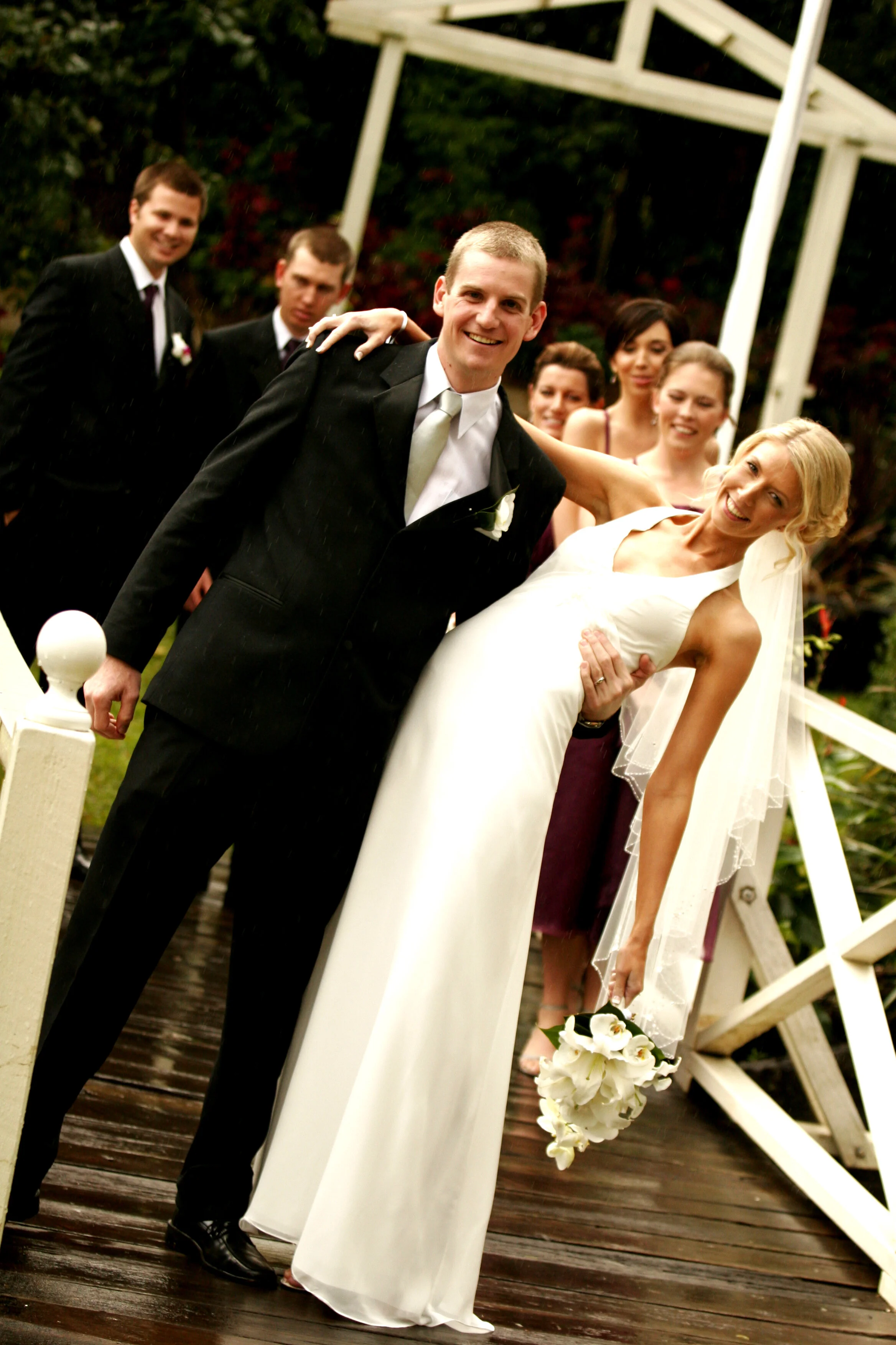 A newlywed couple poses for a photo with their wedding party outdoors on rainy weather, with the groom holding the bride as she holds a bouquet of white flowers.