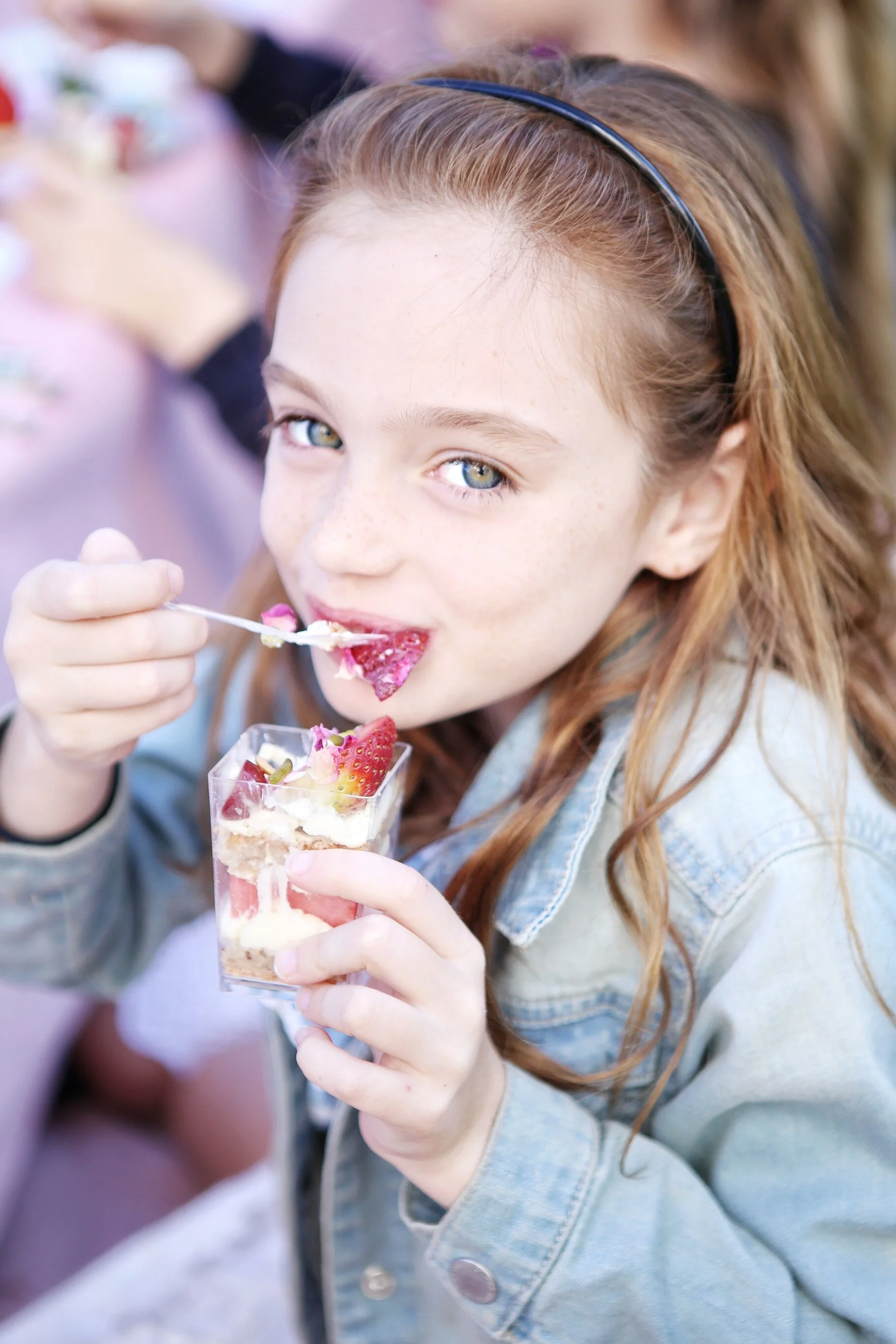 A red-haired girl with freckles and a black headband enjoys eating a strawberry dessert in a clear glass at a gathering.