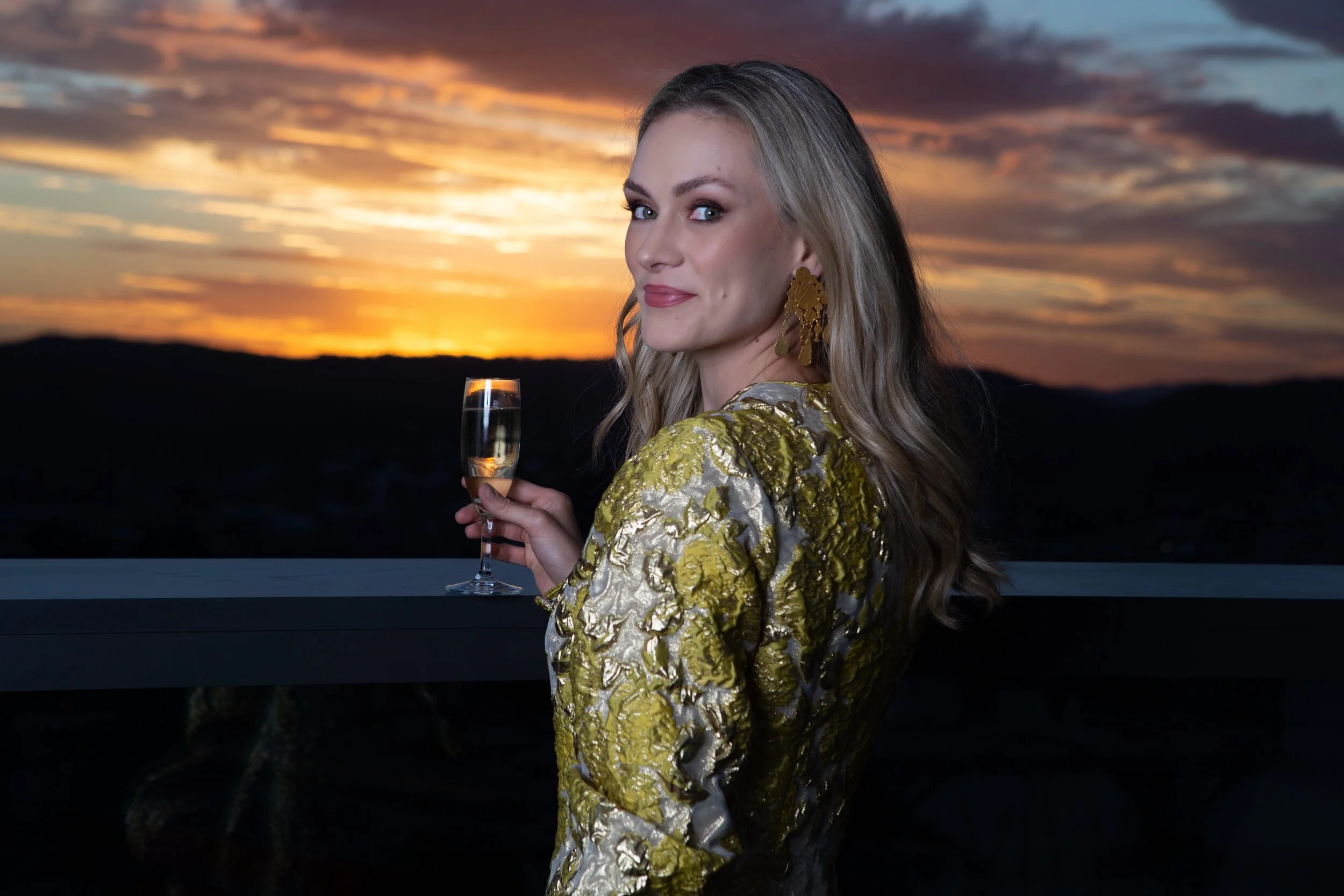 A woman with long blonde hair and earrings, wearing a gold and silver patterned dress, holding a glass of champagne, standing on a balcony at sunset with a scenic view of the horizon and colorful sky.