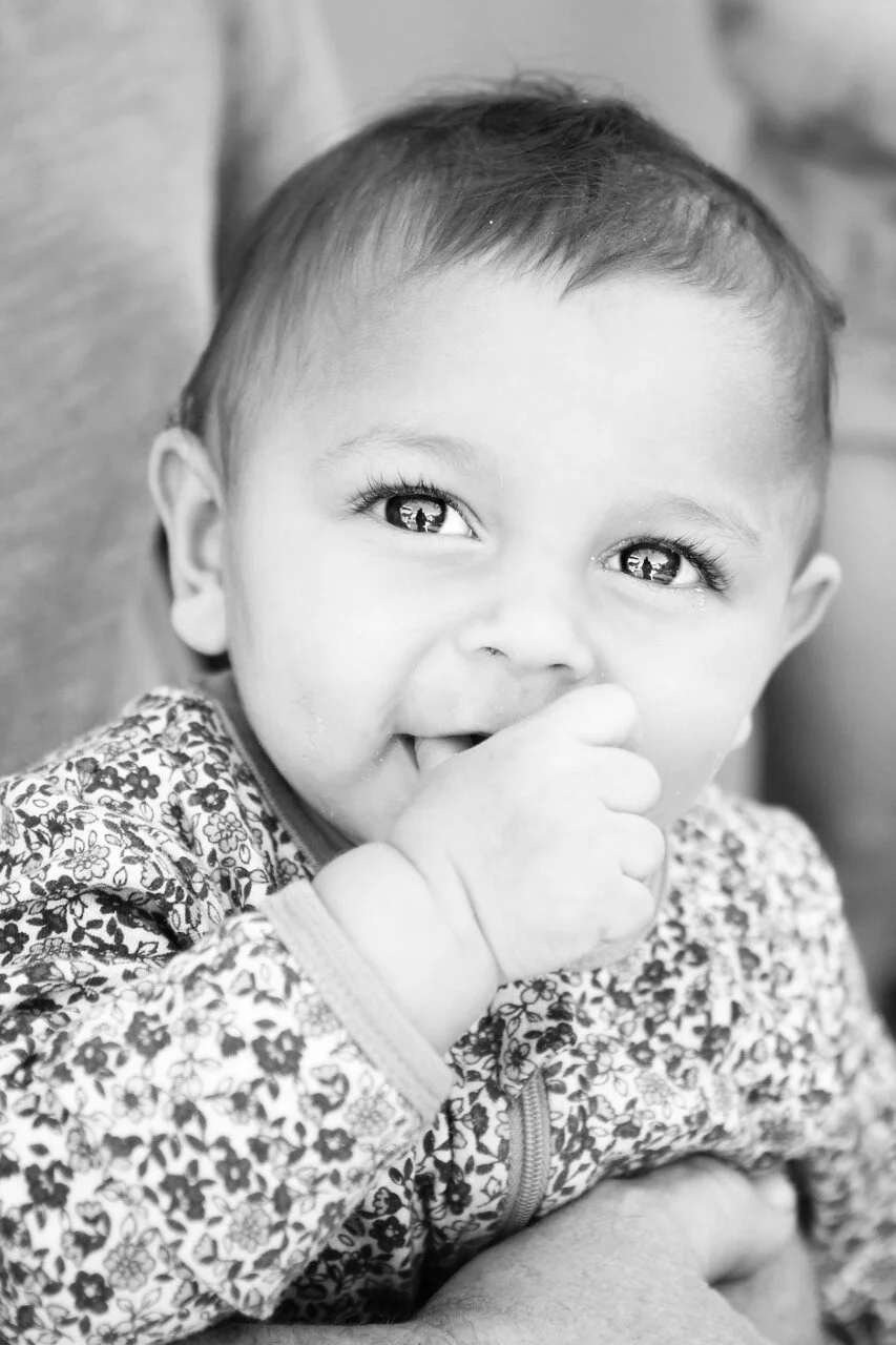 Black and white photo of a baby with big eyes, smiling, with one hand near mouth, wearing a patterned shirt.