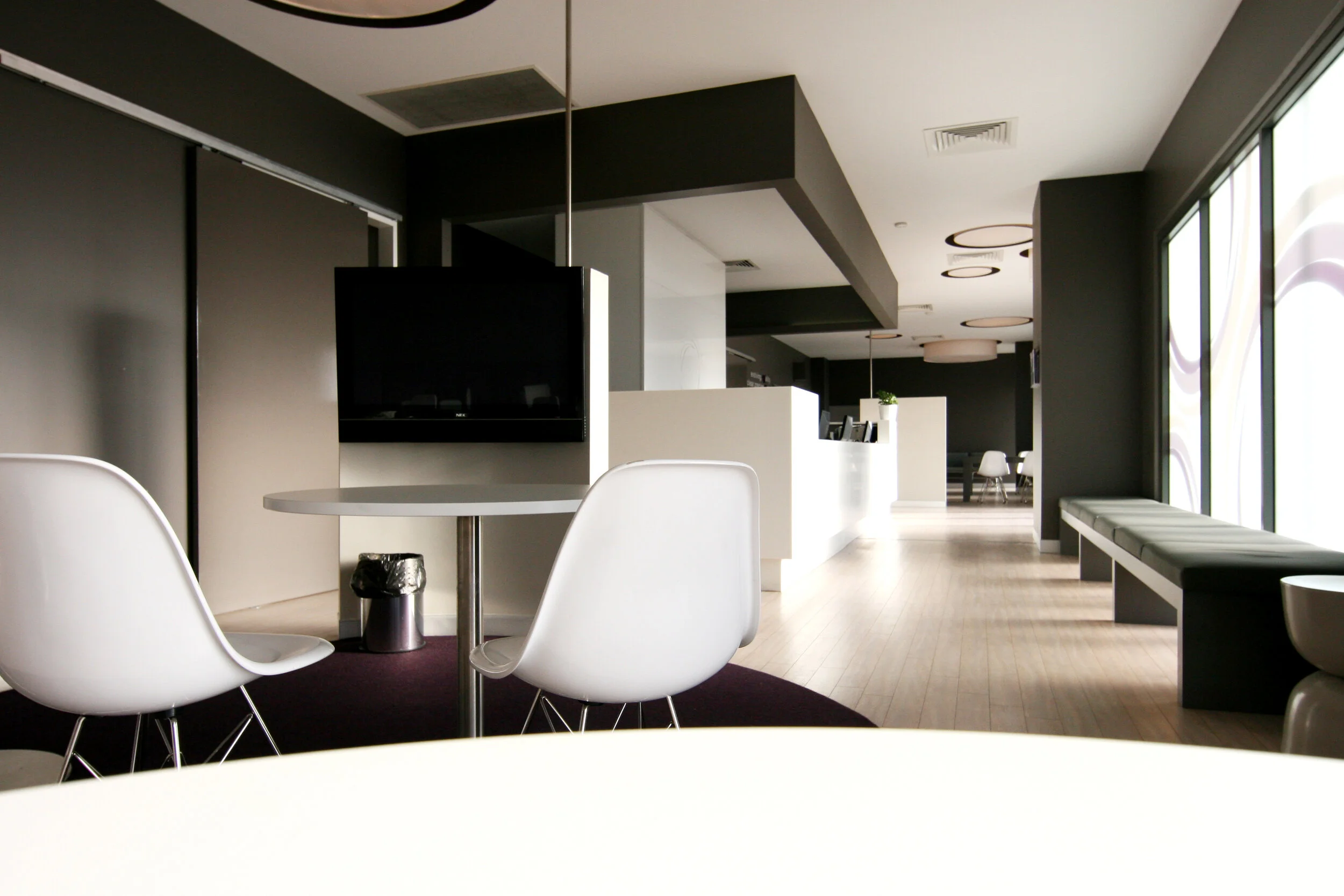 Modern, minimalist interior of a lounge or lobby with white chairs, black and white walls, a flat-screen TV on the wall, large windows, and light wooden floors.