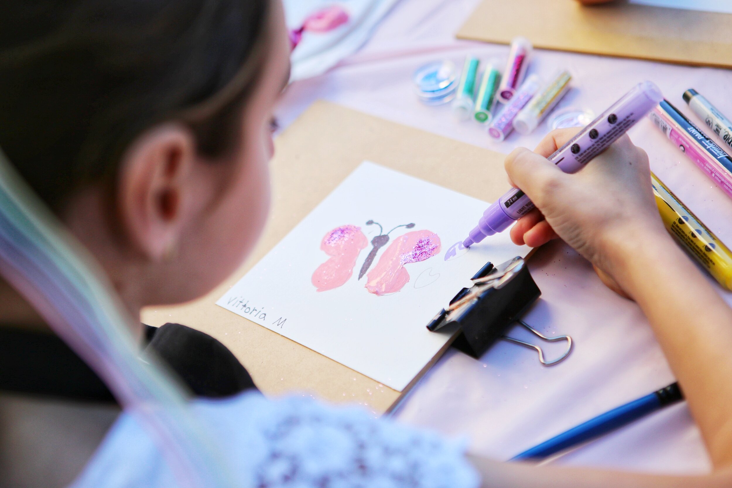 Close-up of a child's hand drawing a butterfly on a piece of paper with purple marker, surrounded by markers and glitter, with the child's head turned away.