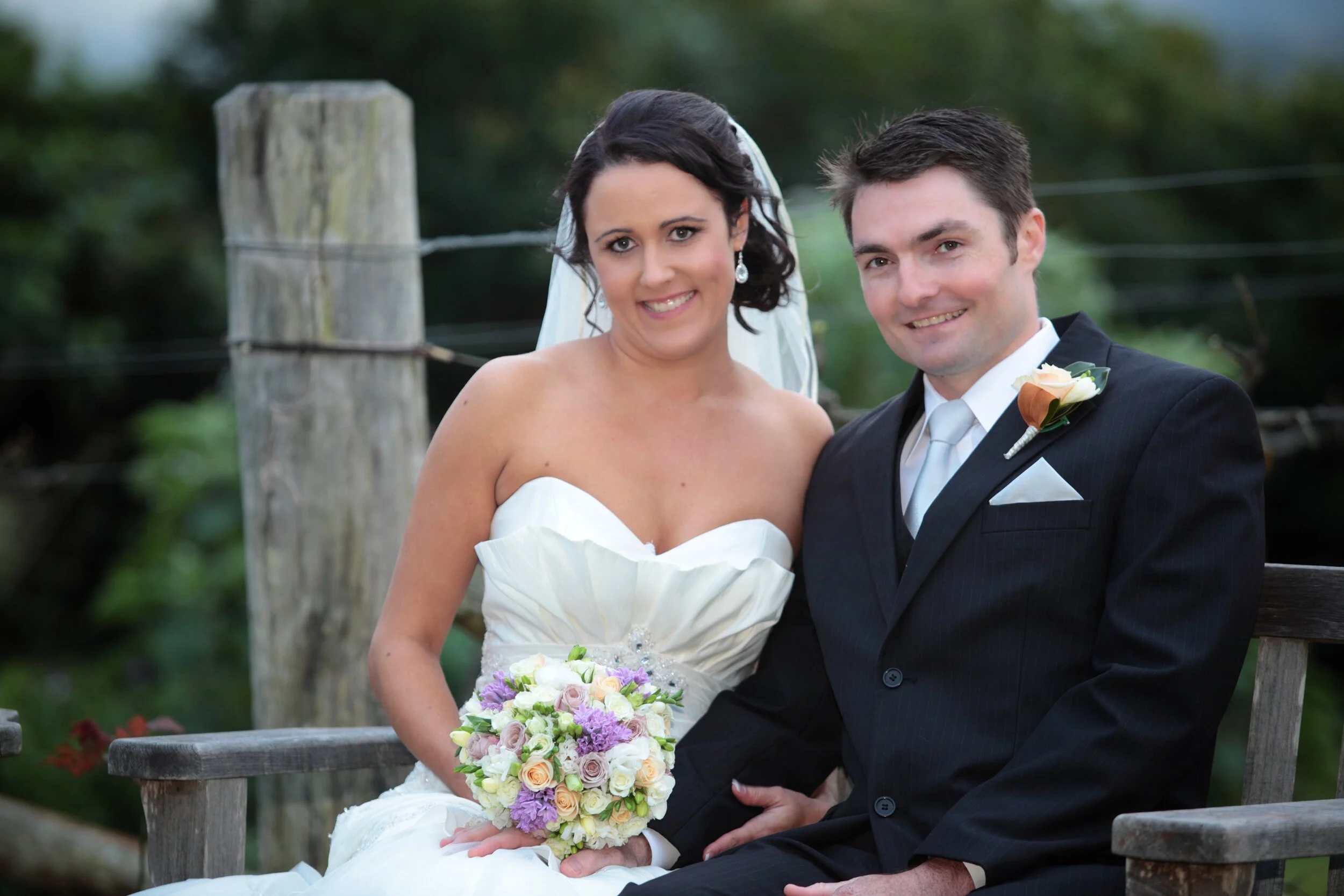 A newlywed couple sitting on a wooden bench outdoors, with the bride holding a bouquet of flowers and both smiling at the camera.