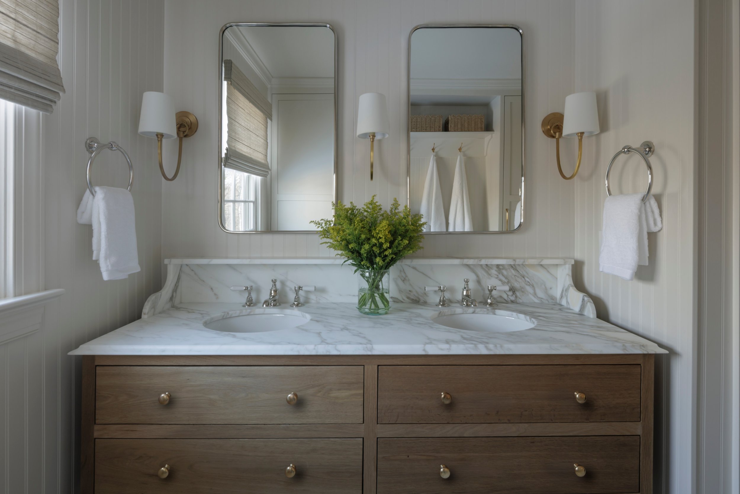 Bathroom vanity with double sinks, marble countertop, two mirrors, wall-mounted light fixtures, towels, and a vase of green flowers.