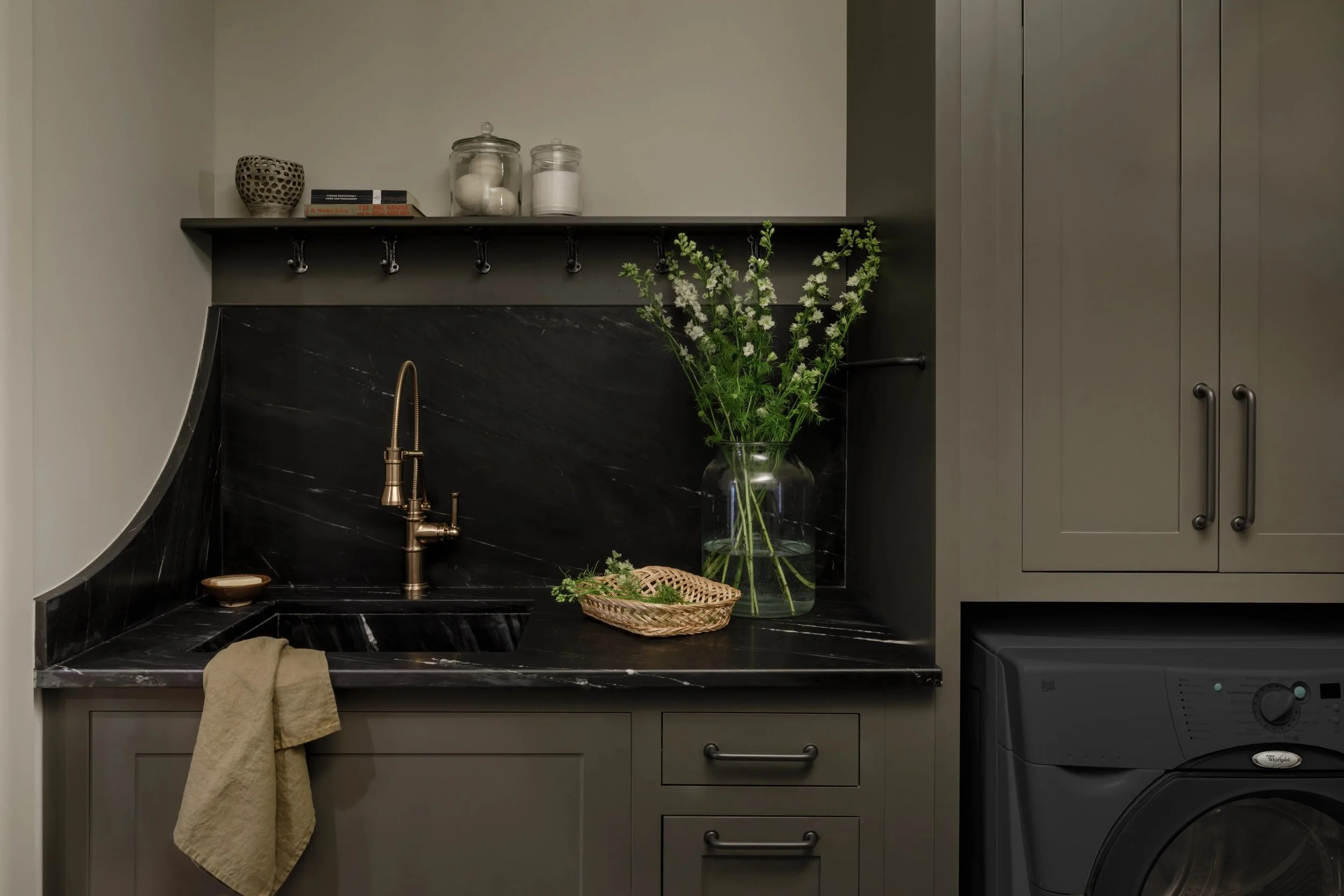 A laundry area with a black marble countertop and backsplash, a brass faucet, a soap dish, a beige towel hanging from the cabinet, a large glass vase with white flowers and greenery, a woven basket, a dark gray washing machine, gray cabinets, and a d