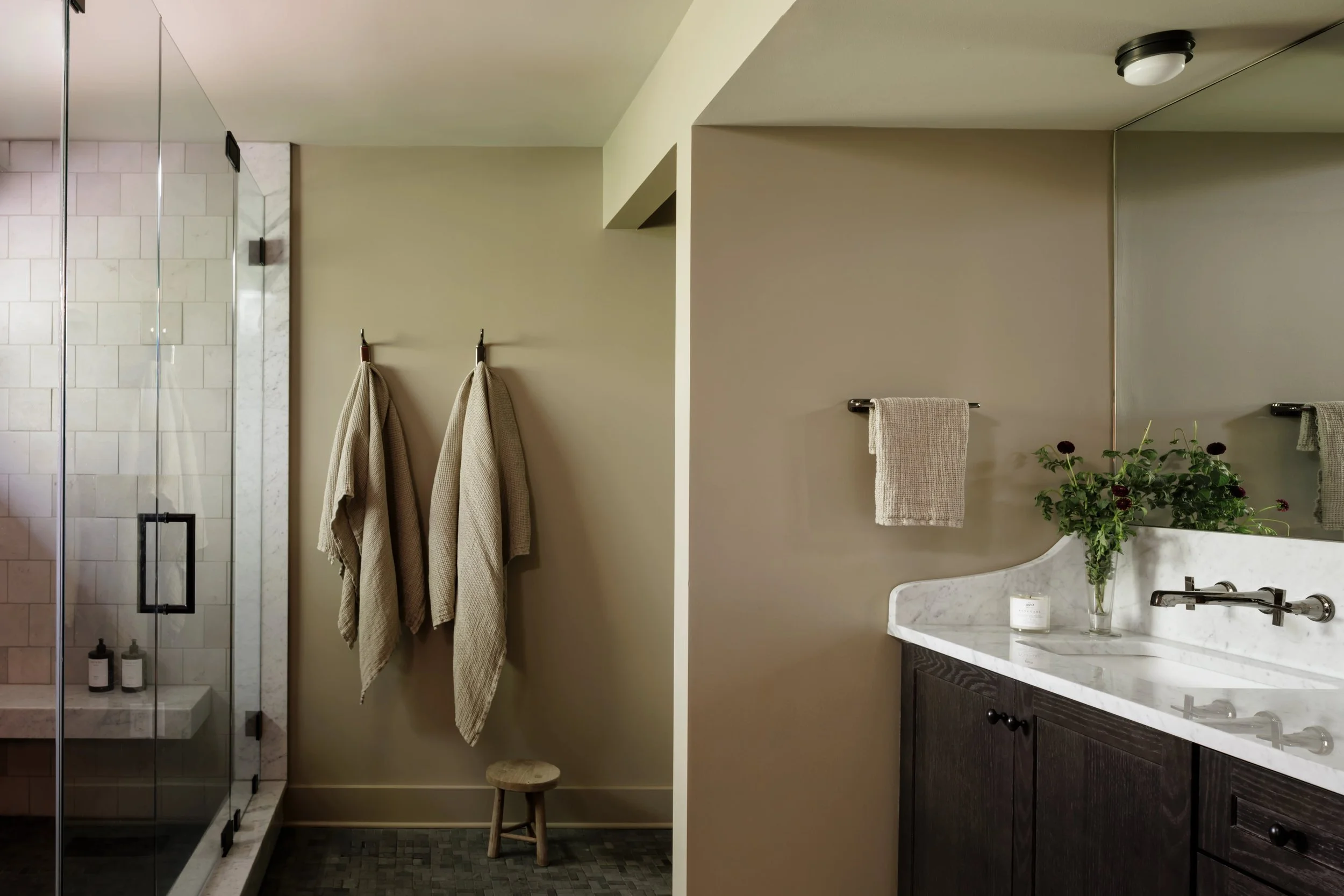 A bathroom with a glass shower enclosure on the left, beige towels hanging on black hooks, a towel bar with a beige towel, a small stool, and a marble countertop with a vase of dark red flowers and a candle.