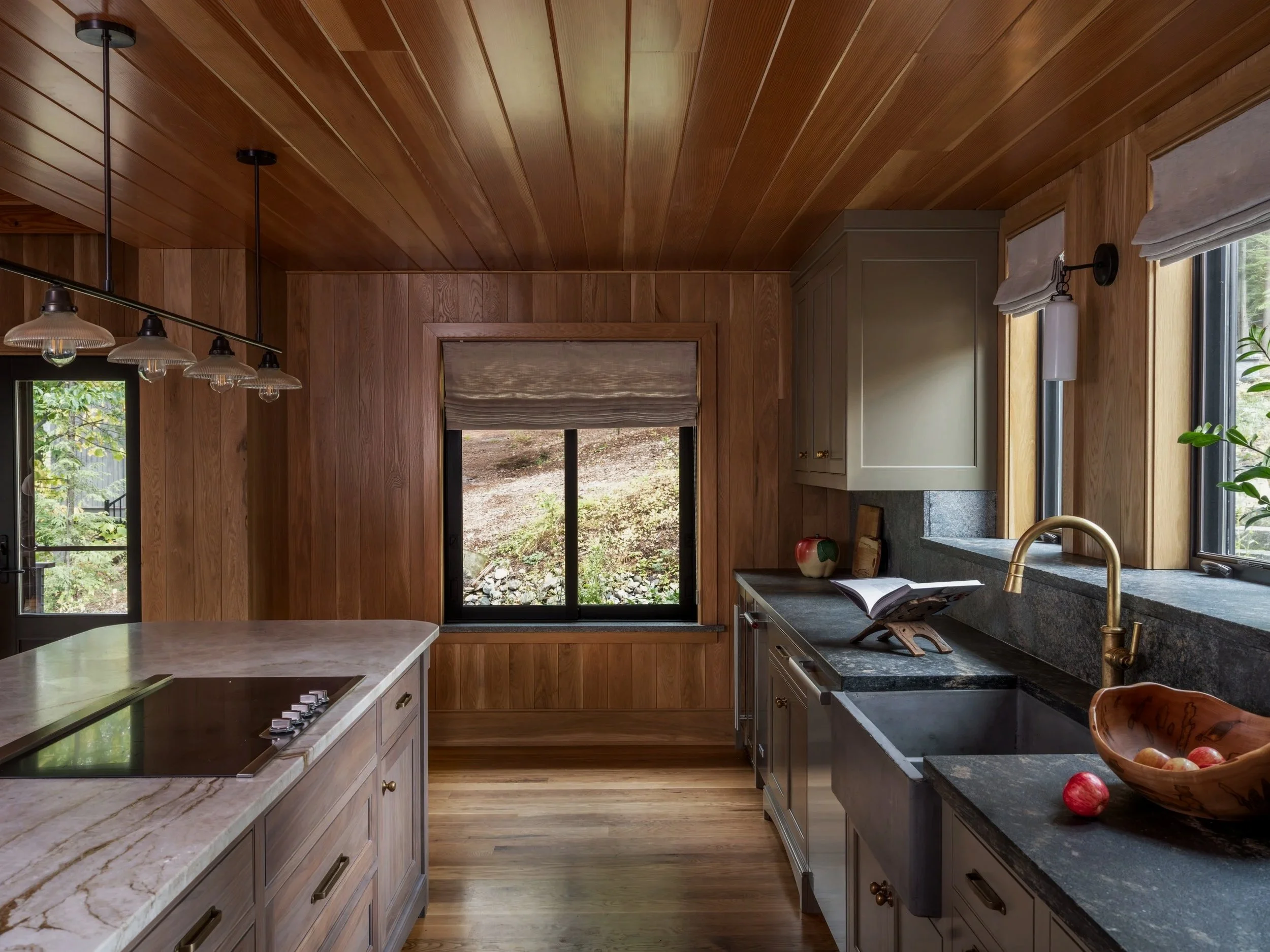 Kitchen with wooden walls and ceiling, black granite countertops, white cabinets, a gold faucet, a large window, a sunny outdoor view, utensils, a bowl of apples, and a book on a stand.