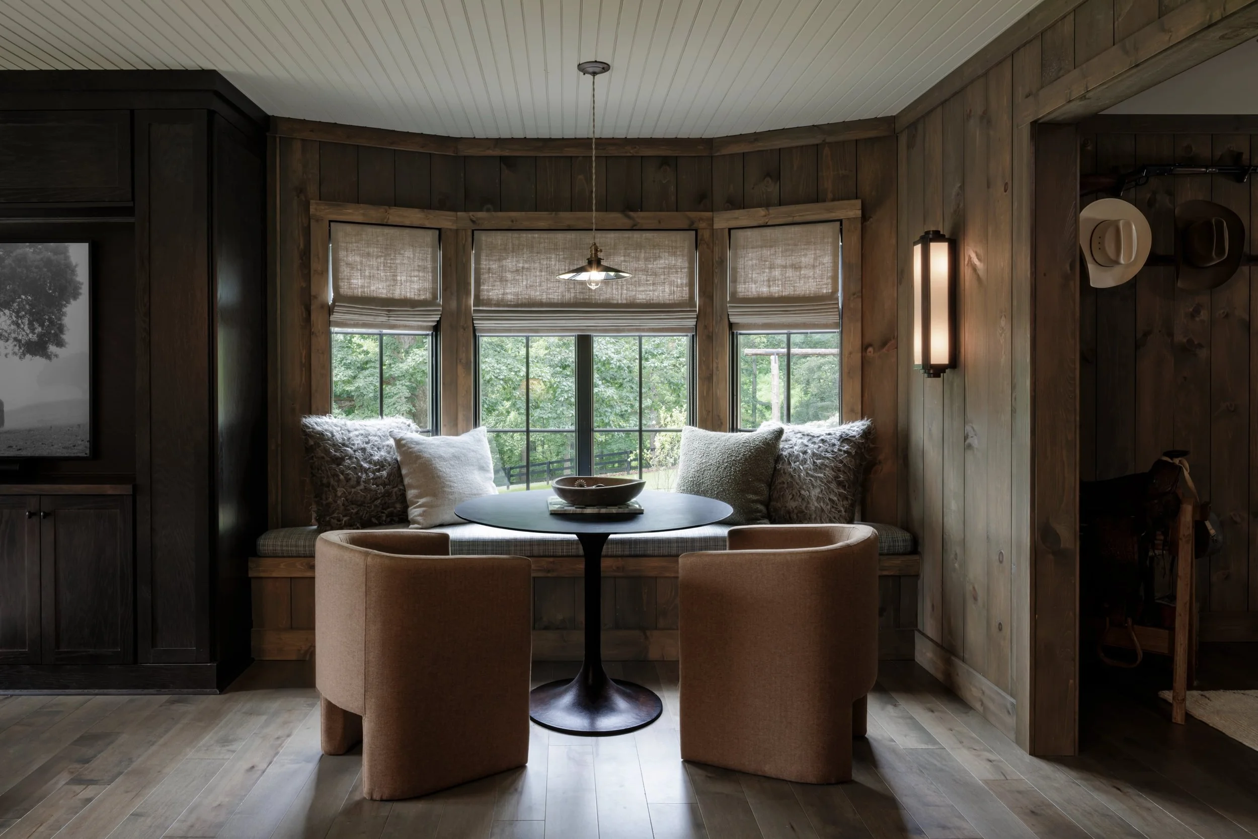 Cozy dining nook with a round table, beige chairs, and a window seat with pillows, surrounded by wood-paneled walls and large windows showing greenery outside.