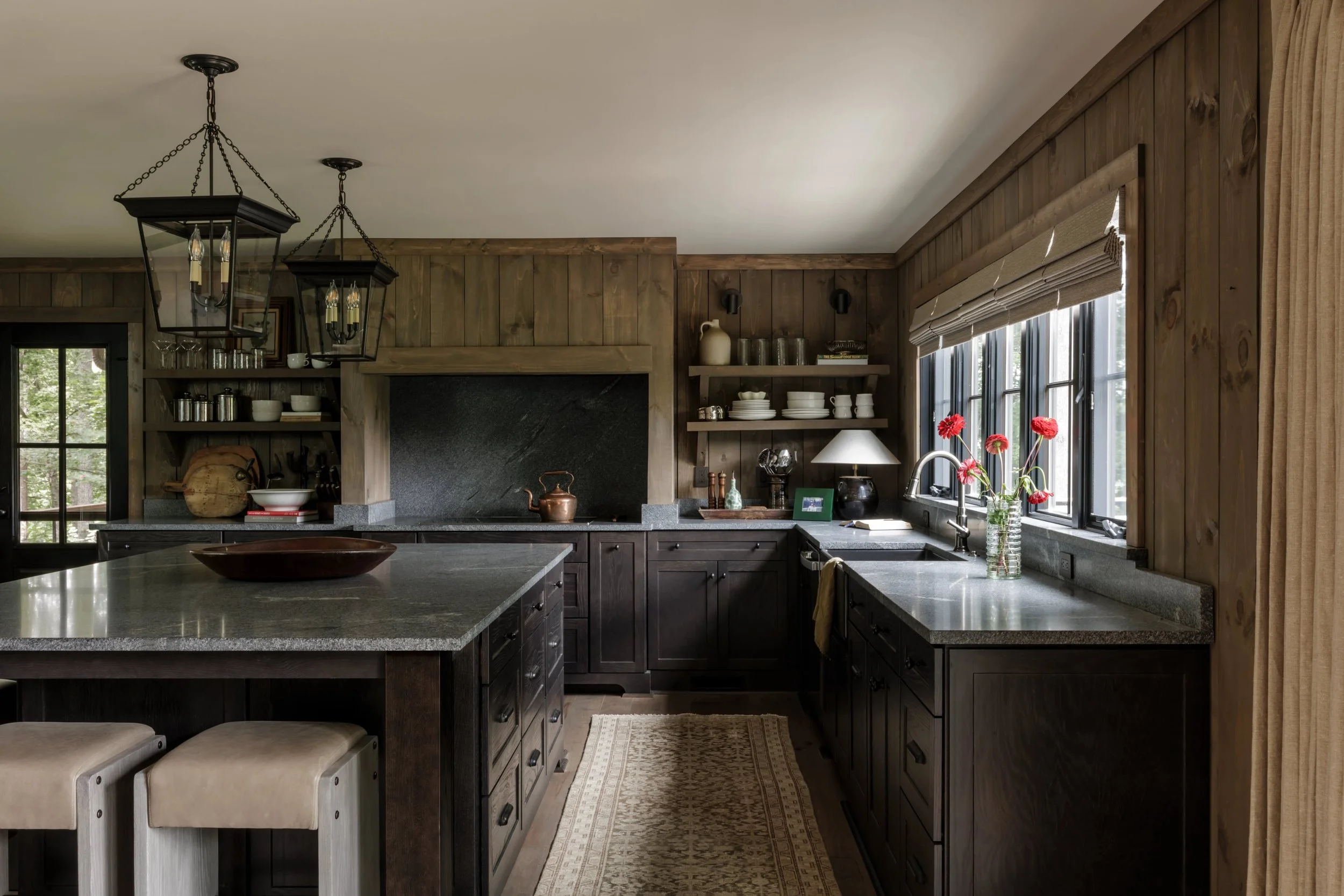 Rustic kitchen with dark wood cabinets, granite countertops, open shelving with dishes, a window with blinds, a vase of red flowers, and a cream-colored curtain.