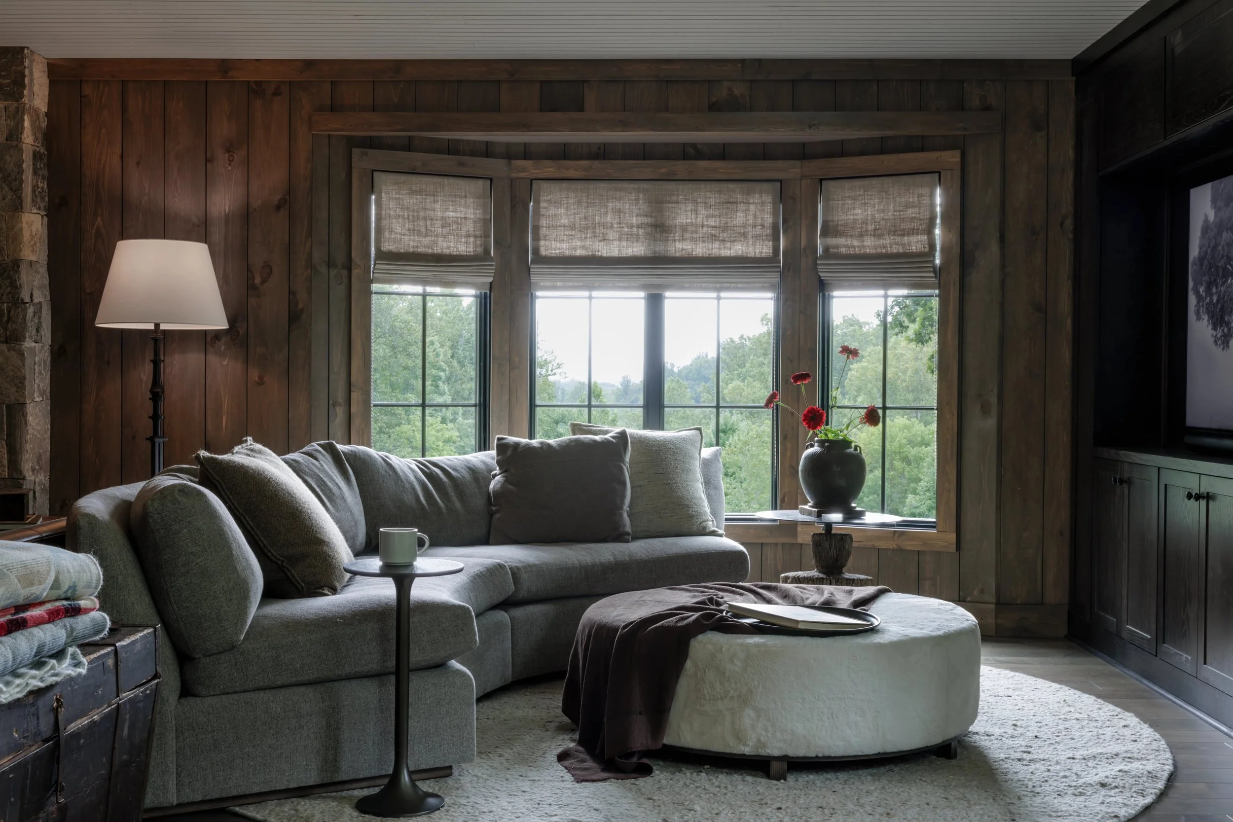 Living room with a gray sectional sofa, a small black side table with a mug, a large ottoman with a book and blanket, a vase of red flowers on a side table, wooden walls, large windows with blinds, and a TV on the wall.