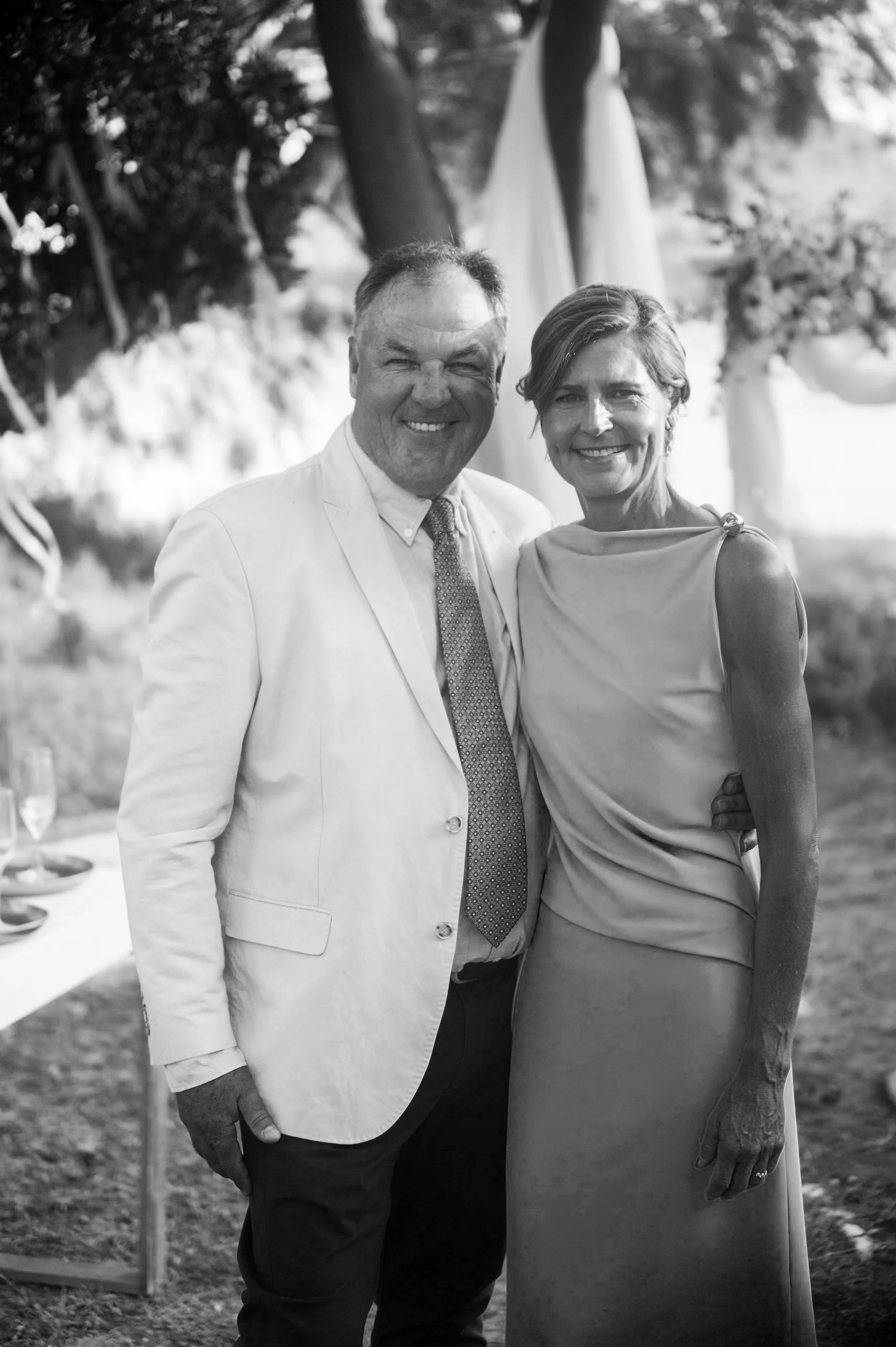 A black and white photo of a smiling man and woman standing close together outdoors. The man is wearing a light-colored suit jacket, shirt, and tie, and the woman is wearing a sleeveless dress. There are trees and outdoor drapery in the background.