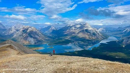 A hiker standing on a the Tent Ridge overlooking the Spray Lake surrounded by mountains under a partly cloudy sky.