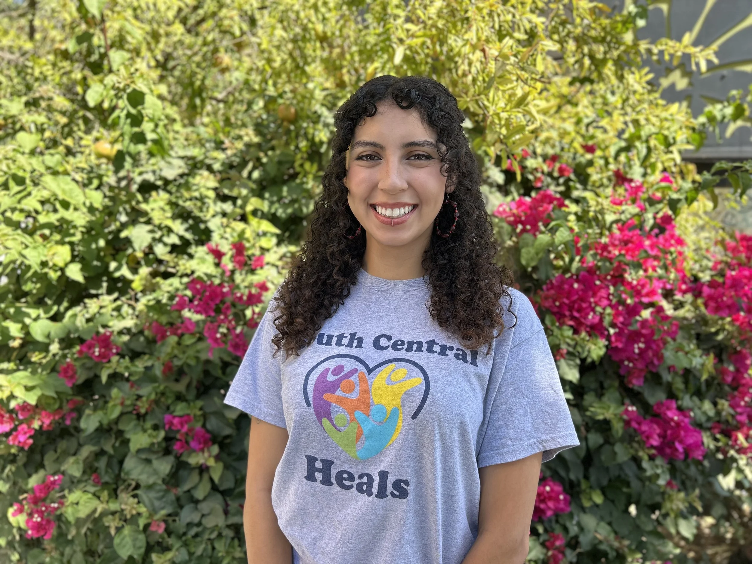 Woman with long curly brown hair, wearing a grey "South Central Heals" shirt smiling at the camera. 
