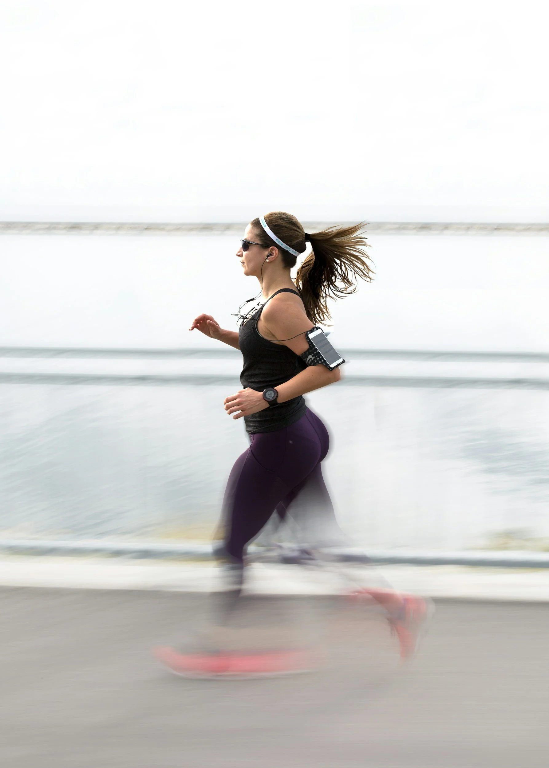 A person in purple spandex leggings and a black tank top wears dark sunglasses, a white hairband, and has their hair tied in a ponytail. They are also  wearing a device on their upper arm with wired headphones. running in a blurred image.