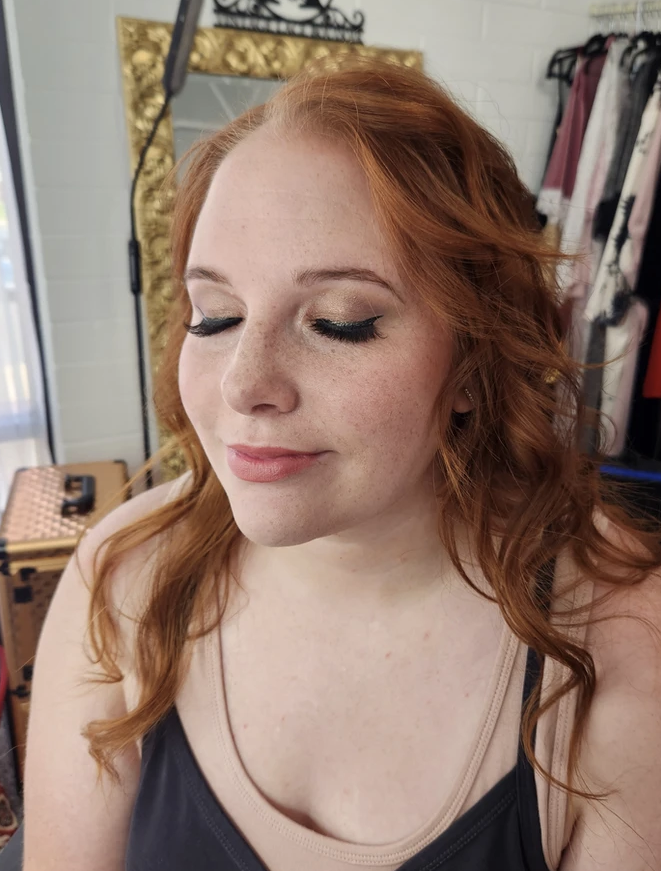 Woman with red hair and closed eyes, wearing makeup, in a room with clothing and a decorative mirror.