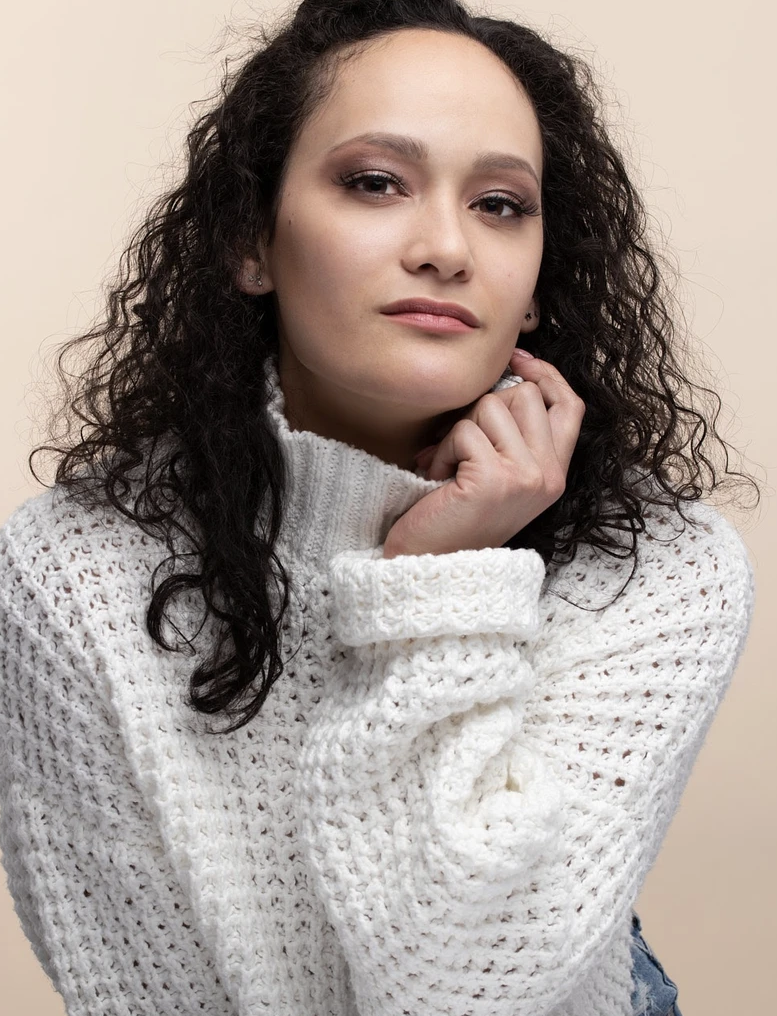 Woman with curly hair wearing a white knit sweater, posing with hand on chin against a beige background.