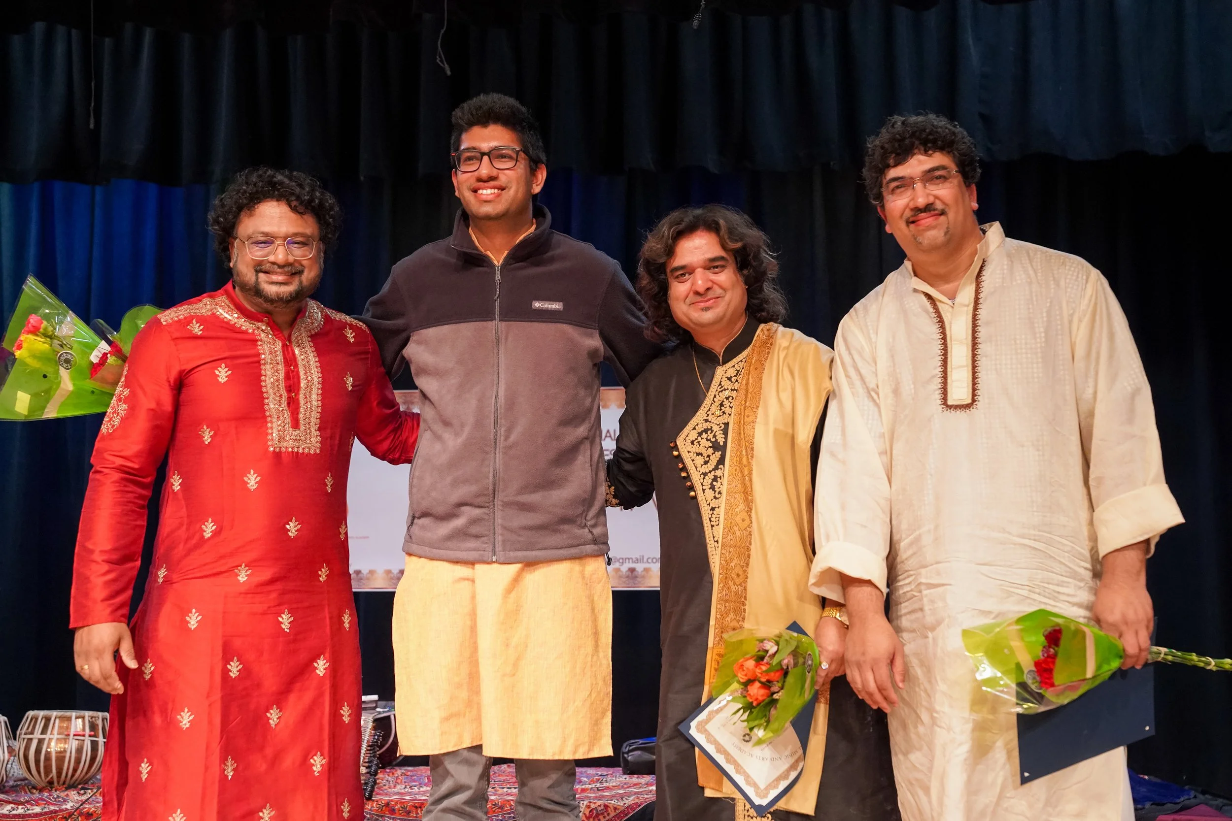 Group of five men standing on stage, some holding bouquets of flowers, dressed in traditional and casual Indian clothing, smiling for a photo.