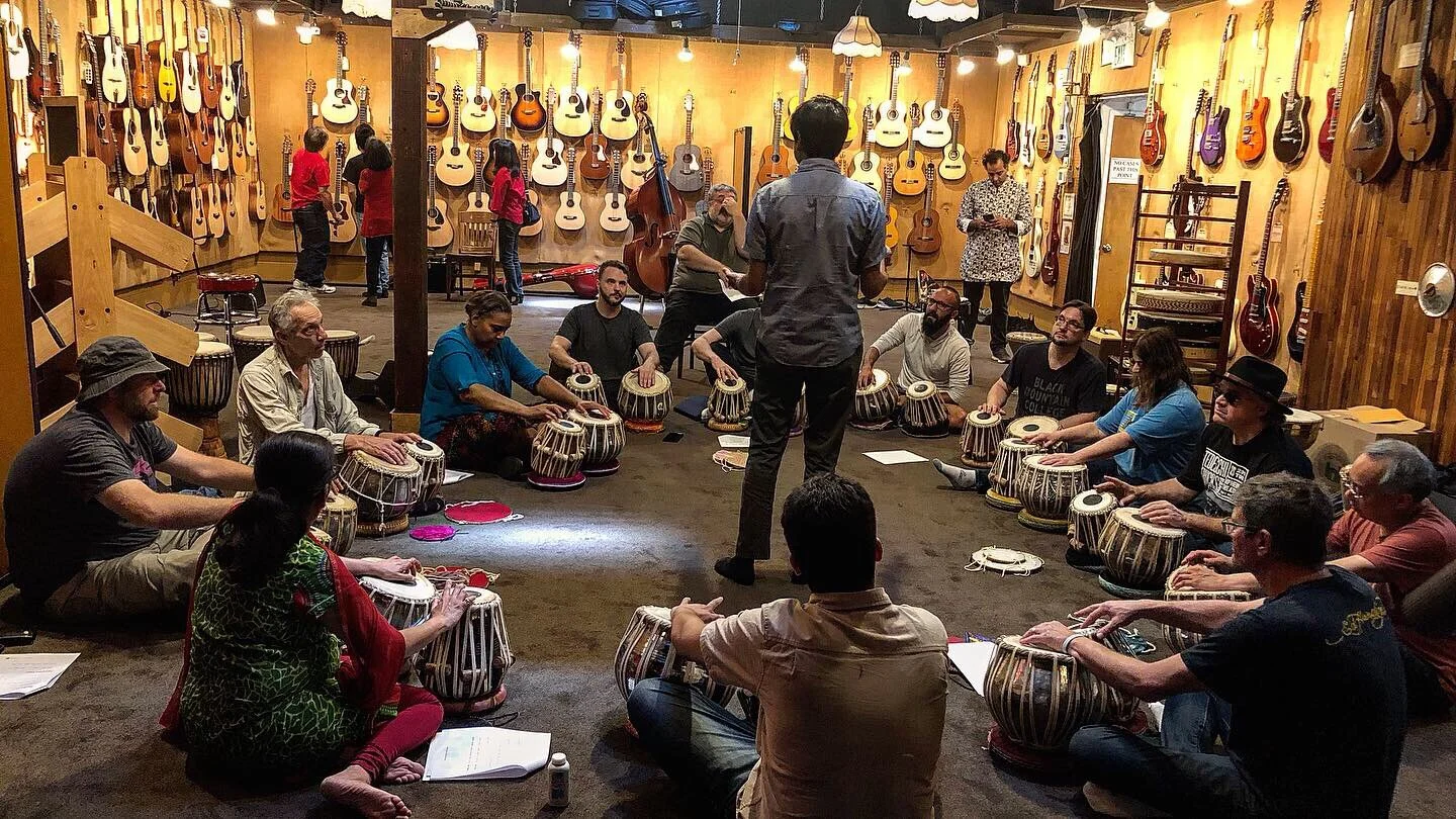 A group of people sits in a circle, playing traditional drums during a music class or workshop inside a store with guitars on the walls.