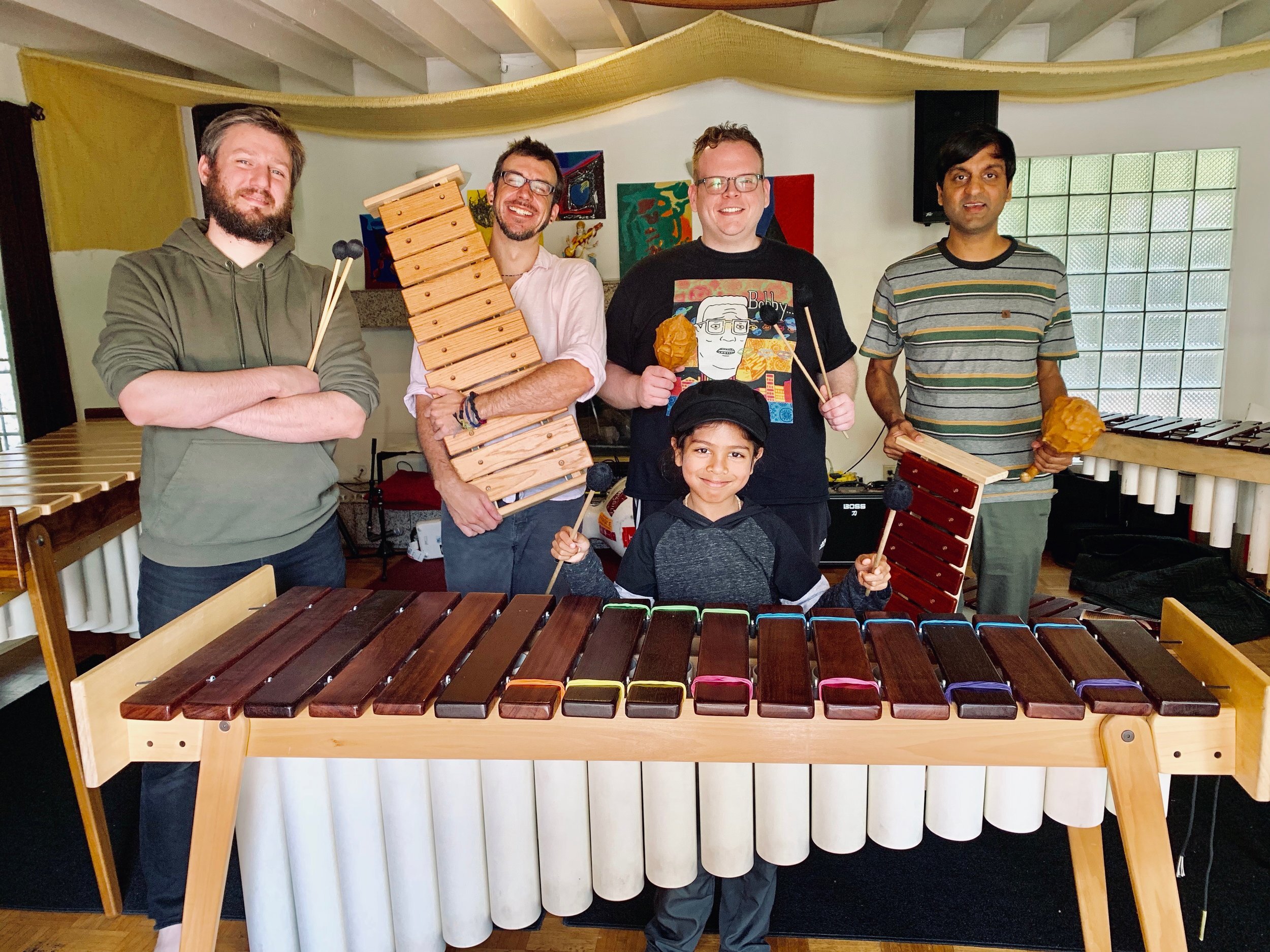Five adults and one child posing with and holding various colorful percussion instruments, including xylophones and maracas, in a room with yellow drapes and artwork on the walls.