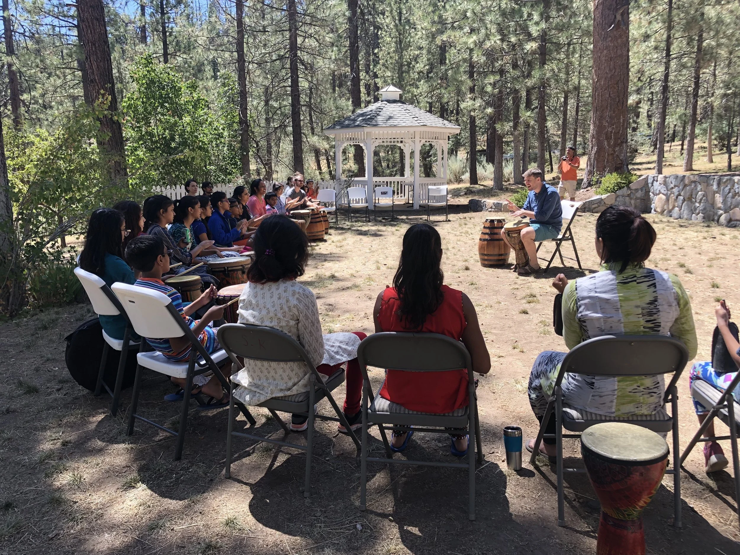 Group of children and adults attending an outdoor drumming and music class in a wooded area with a white gazebo in the background.