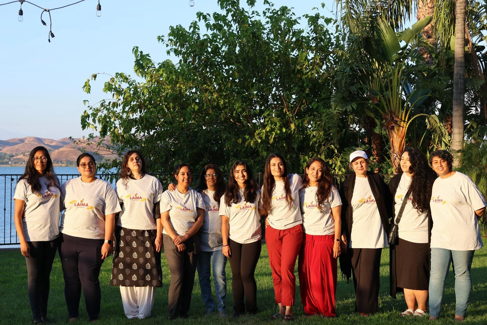 A group of eleven women standing outdoors on grass, posing for a photo with a large tree and a body of water in the background. They are wearing matching white T-shirts with a logo that says "LAIMA."