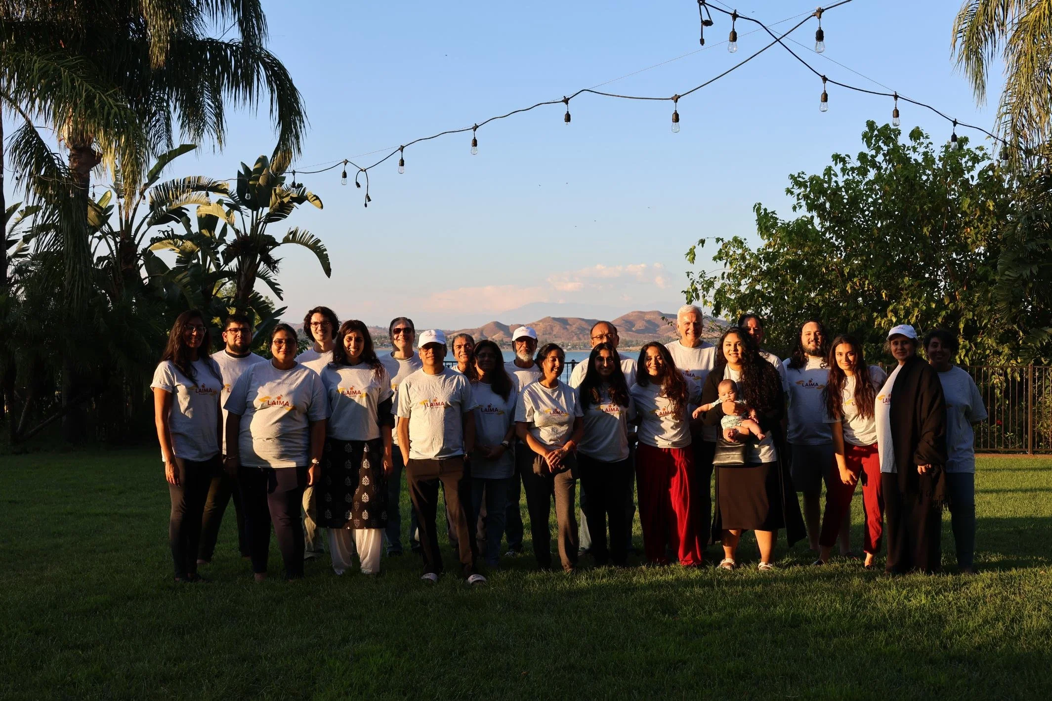 Group of people standing outdoors on grass, with trees and mountains in the background under string lights, during sunset.