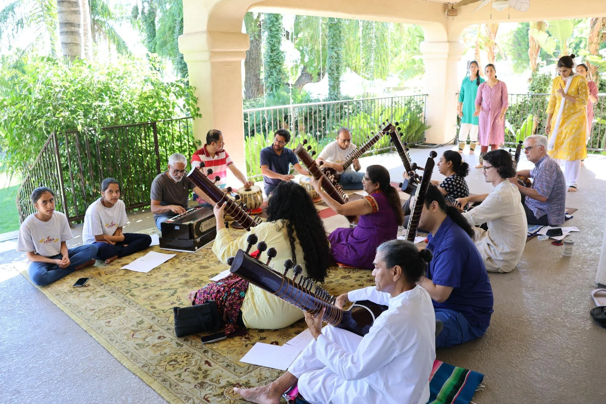 A group of people seated on a rug playing traditional Indian musical instruments called veenas, with some individuals in front sitting cross-legged. Other people are sitting on the porch observing or participating, some standing and walking across th
