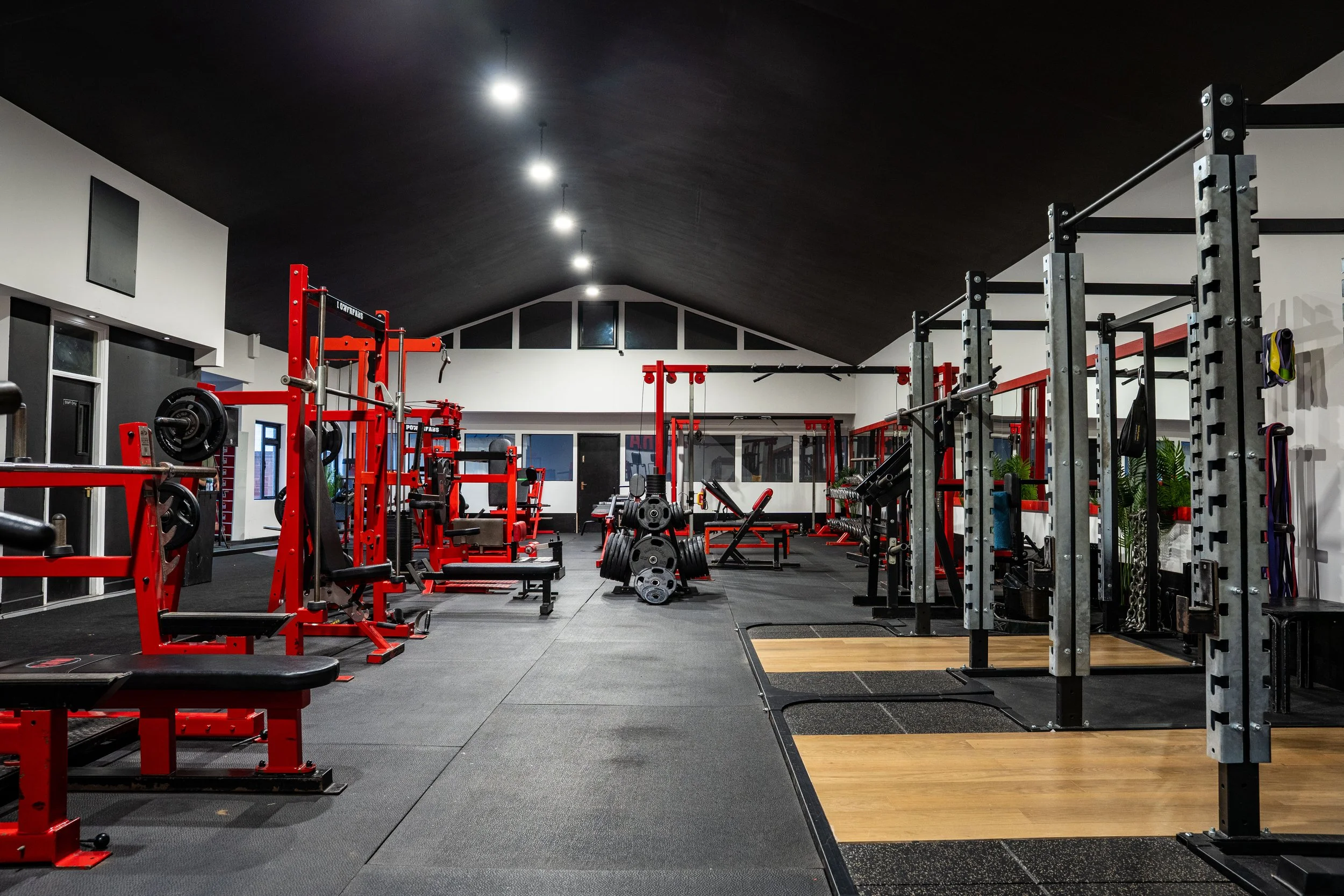 Empty gym with various red and black workout equipment including weights, benches, and squat racks, illuminated by ceiling lights.