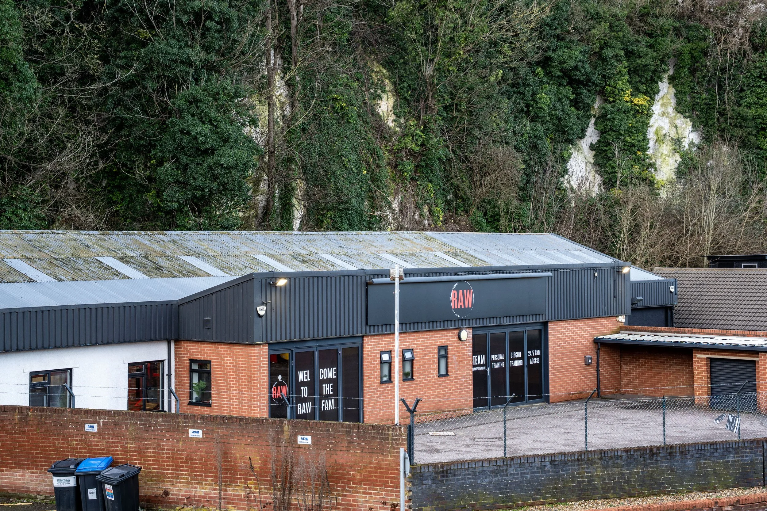 An industrial building with a red brick exterior and black roofing, surrounded by a brick wall and chain-link fence. The building has large windows and signs welcoming people to the RAW gym, indicating it offers team training, personal training, circ