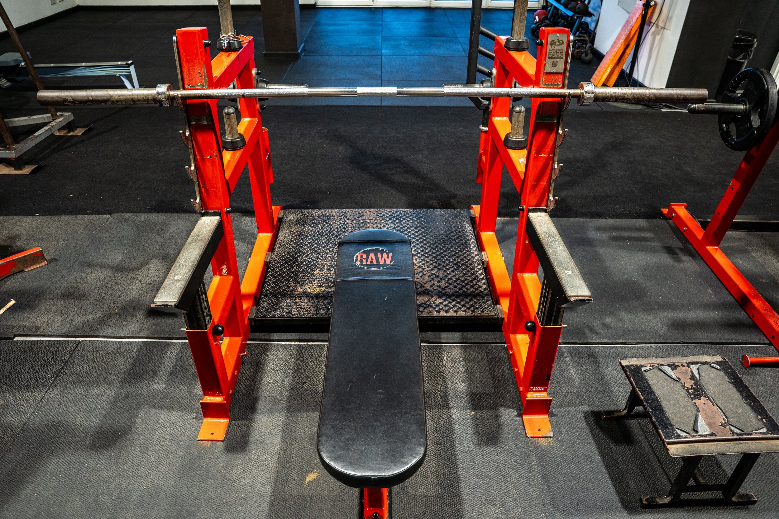 A gym workout station with a loaded barbell, an adjustable bench labeled 'RAW', and a small rusty step platform nearby.