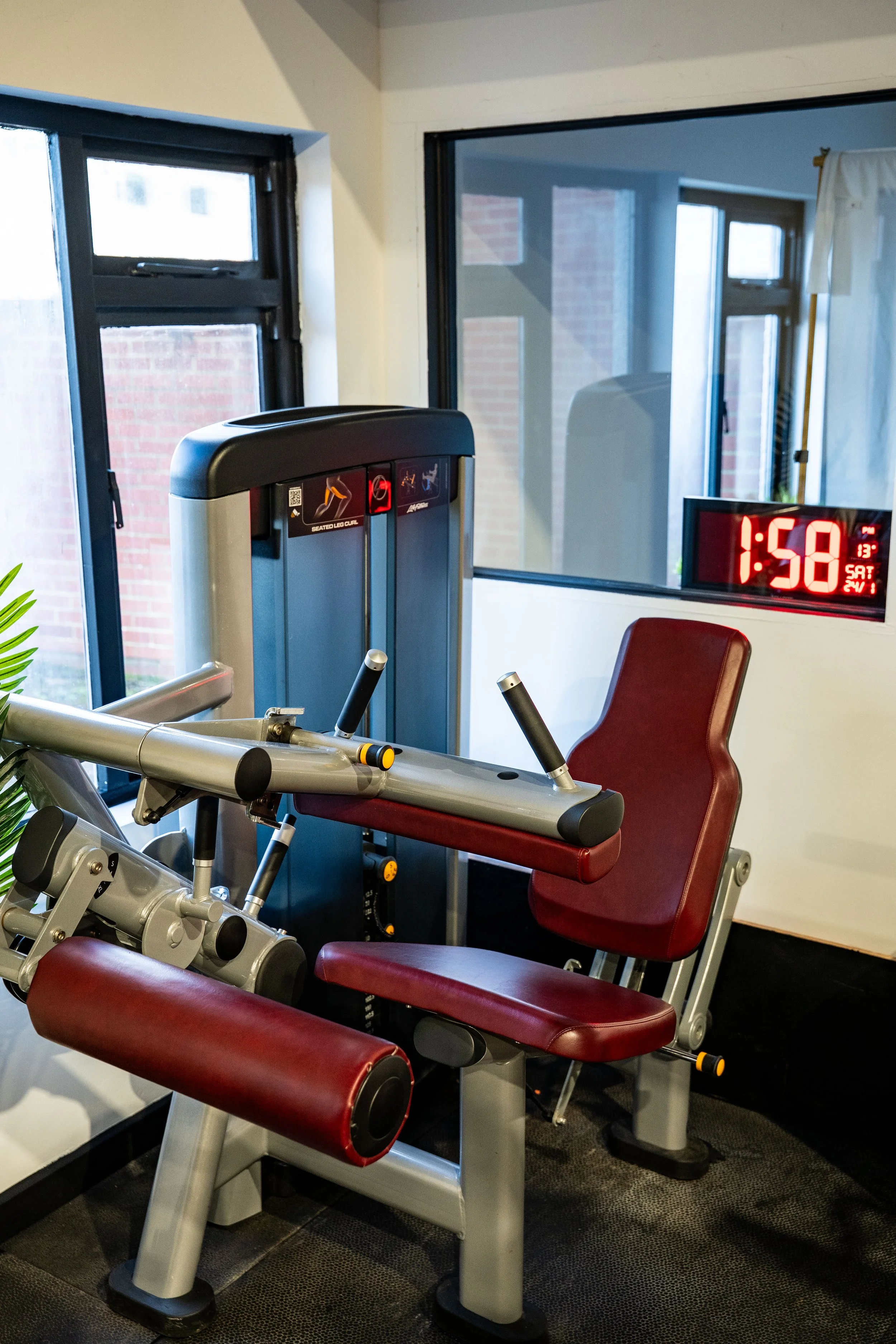 Exercise equipment in a gym, including a seated leg curl machine with red padding, a computer monitor displaying 1:58, and windows in the background.
